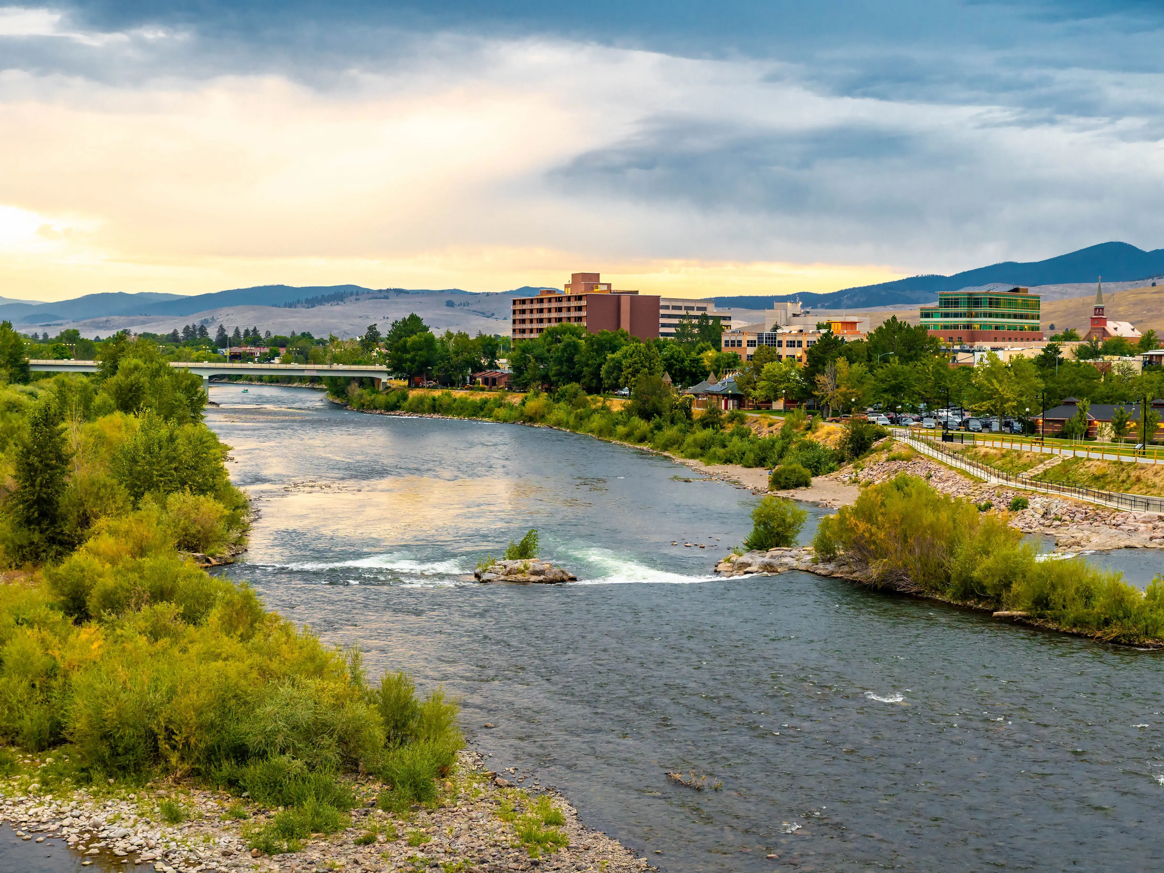 A scenic view of the Clark Fork River winding through downtown Missoula, with city lights beginning to glow as the sun sets