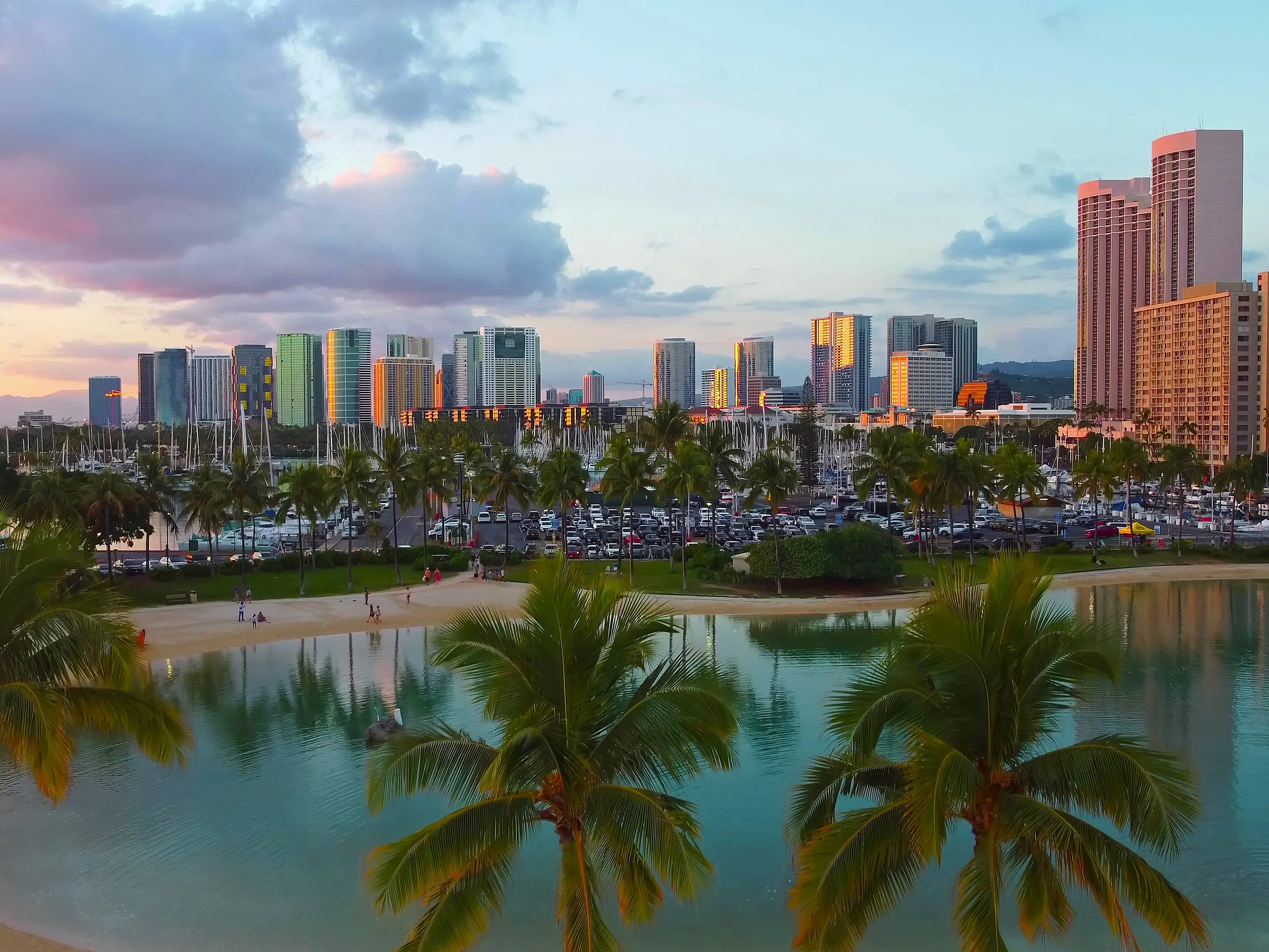 Honolulu skyline and lagoon at sunset. Scenic view of a tropical lagoon with palm trees, sandy beach, and the illuminated Honolulu skyline reflecting the warm hues of sunset