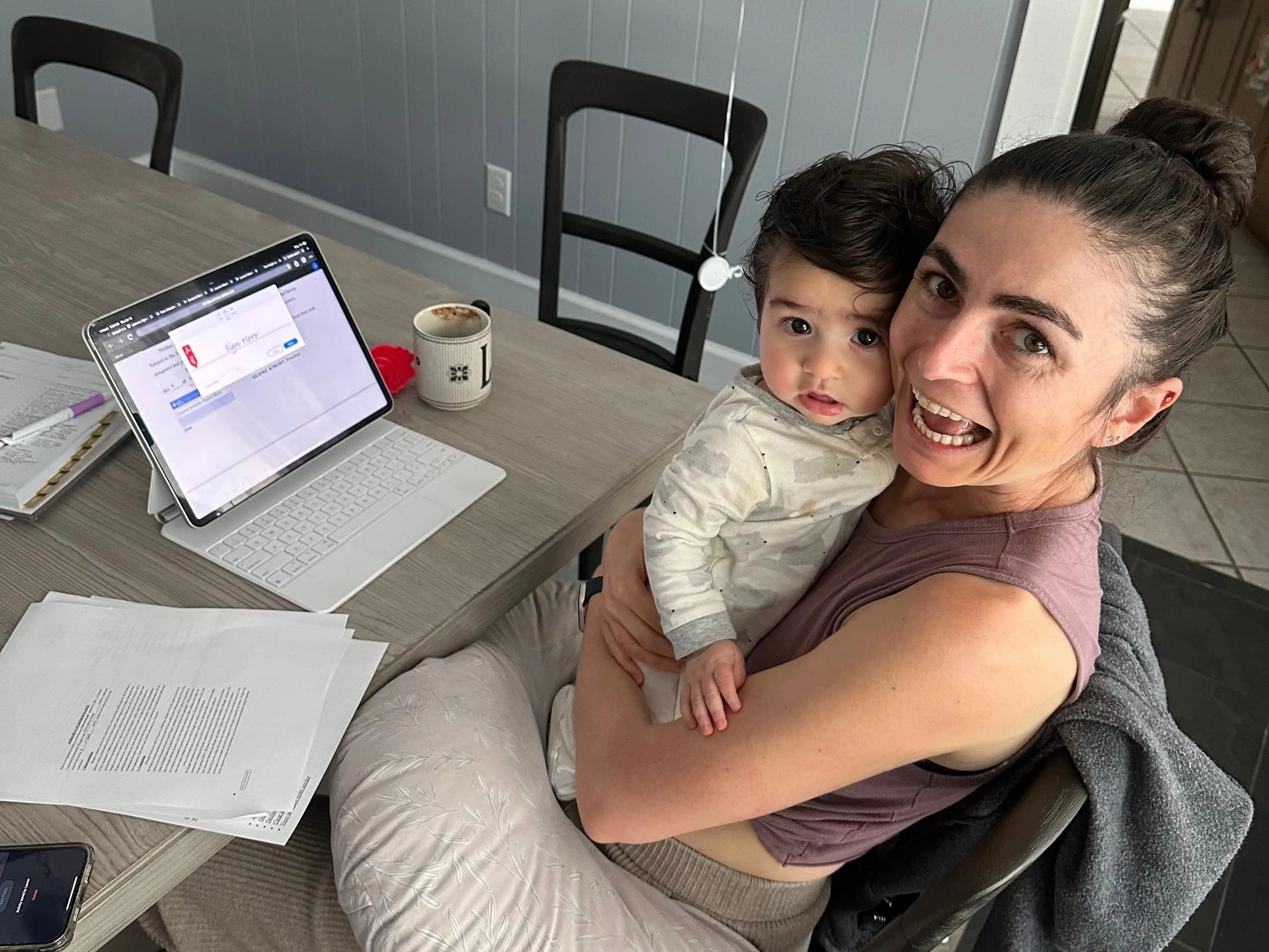The writer holding her child and sitting at a table with papers and a laptop.