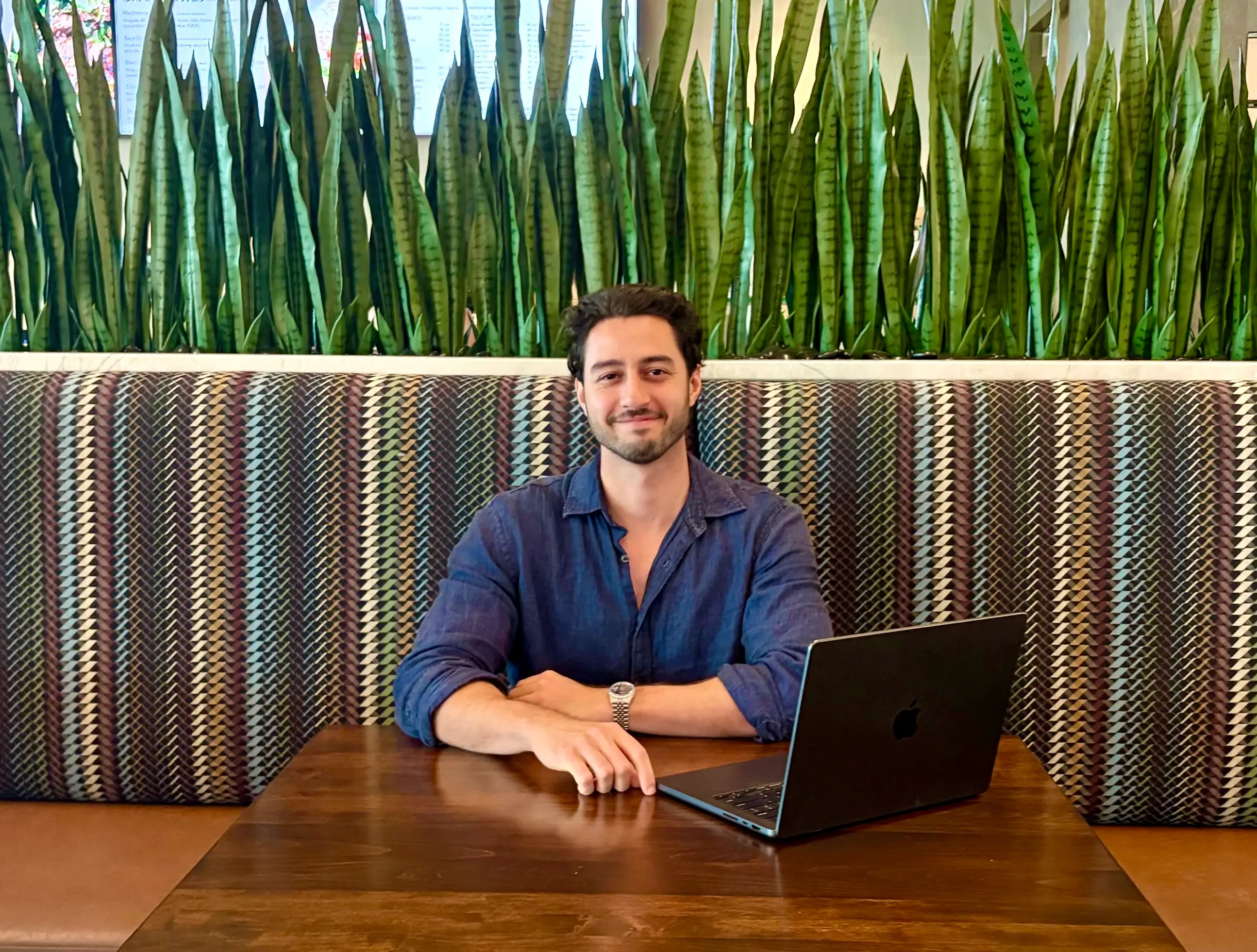 A young man in a button-down shirt sits at a desk with an open laptop.