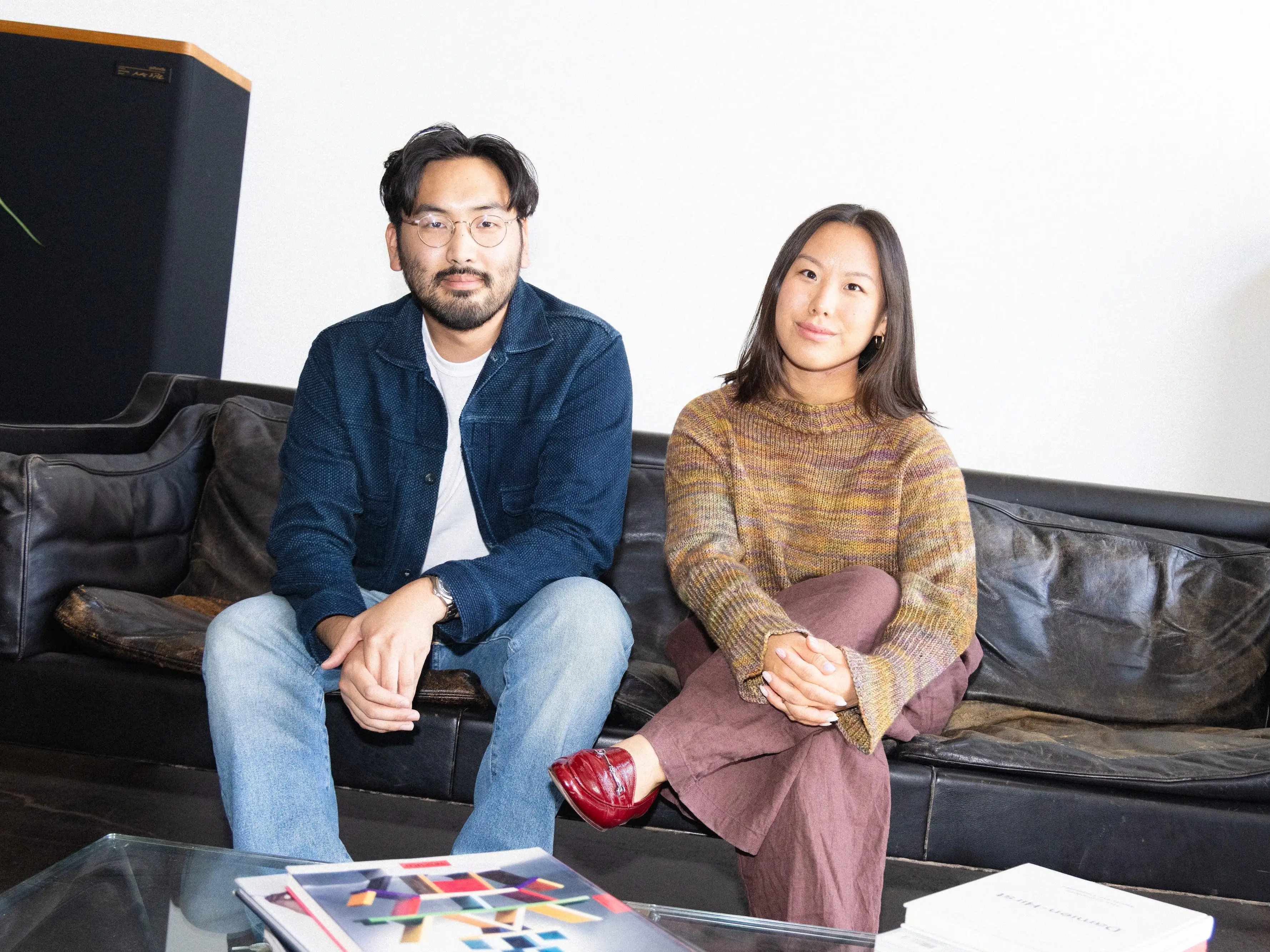 Two people sit on a black leather couch in a bright room with a glass coffee table in front of them.