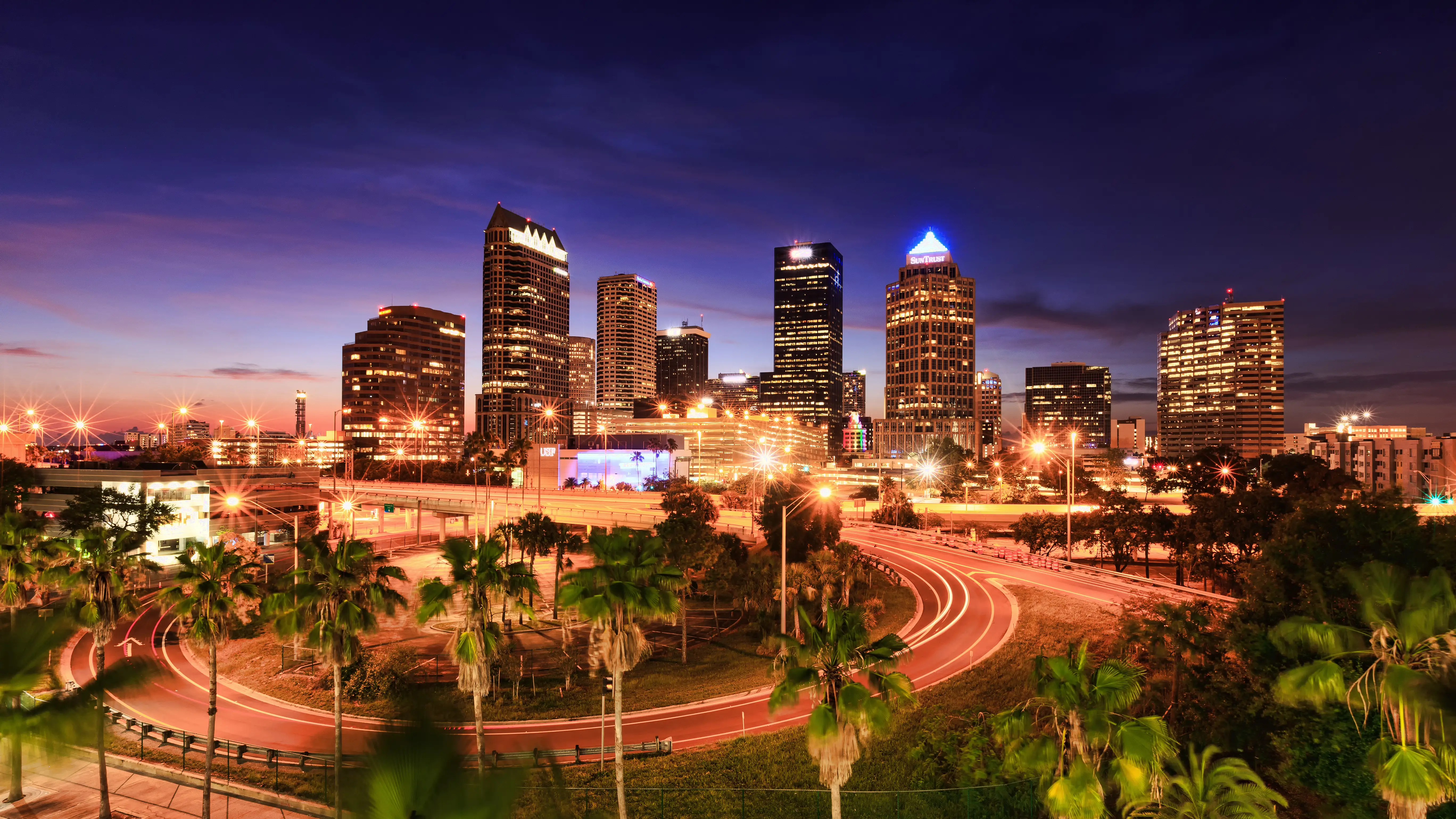 Downtown skyline glows at dusk with illuminated high-rises, palm trees, and curving highway light trails.