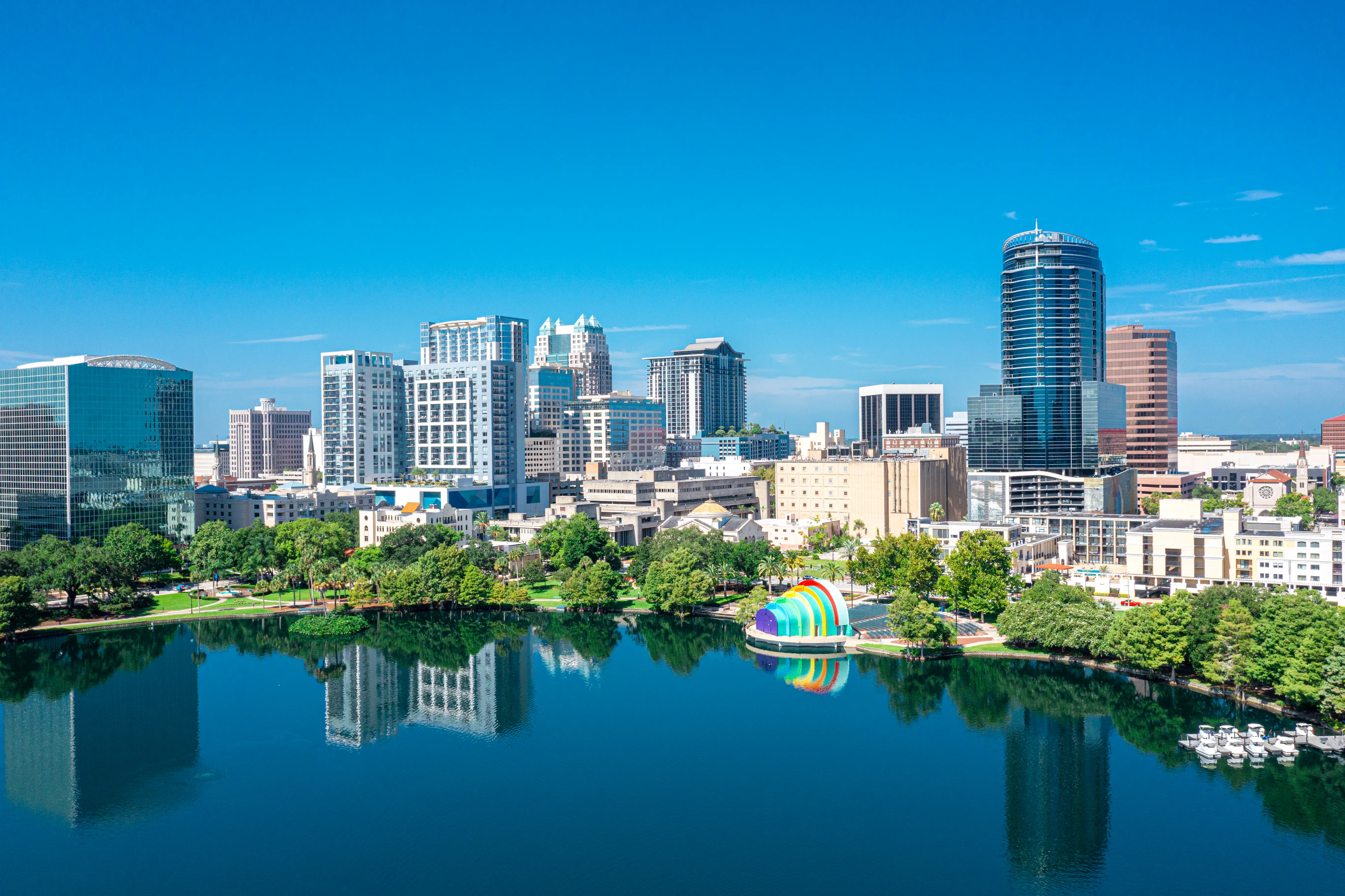 Downtown Orlando skyline rises behind Lake Eola Park with building reflections and a colorful lakeside amphitheater.