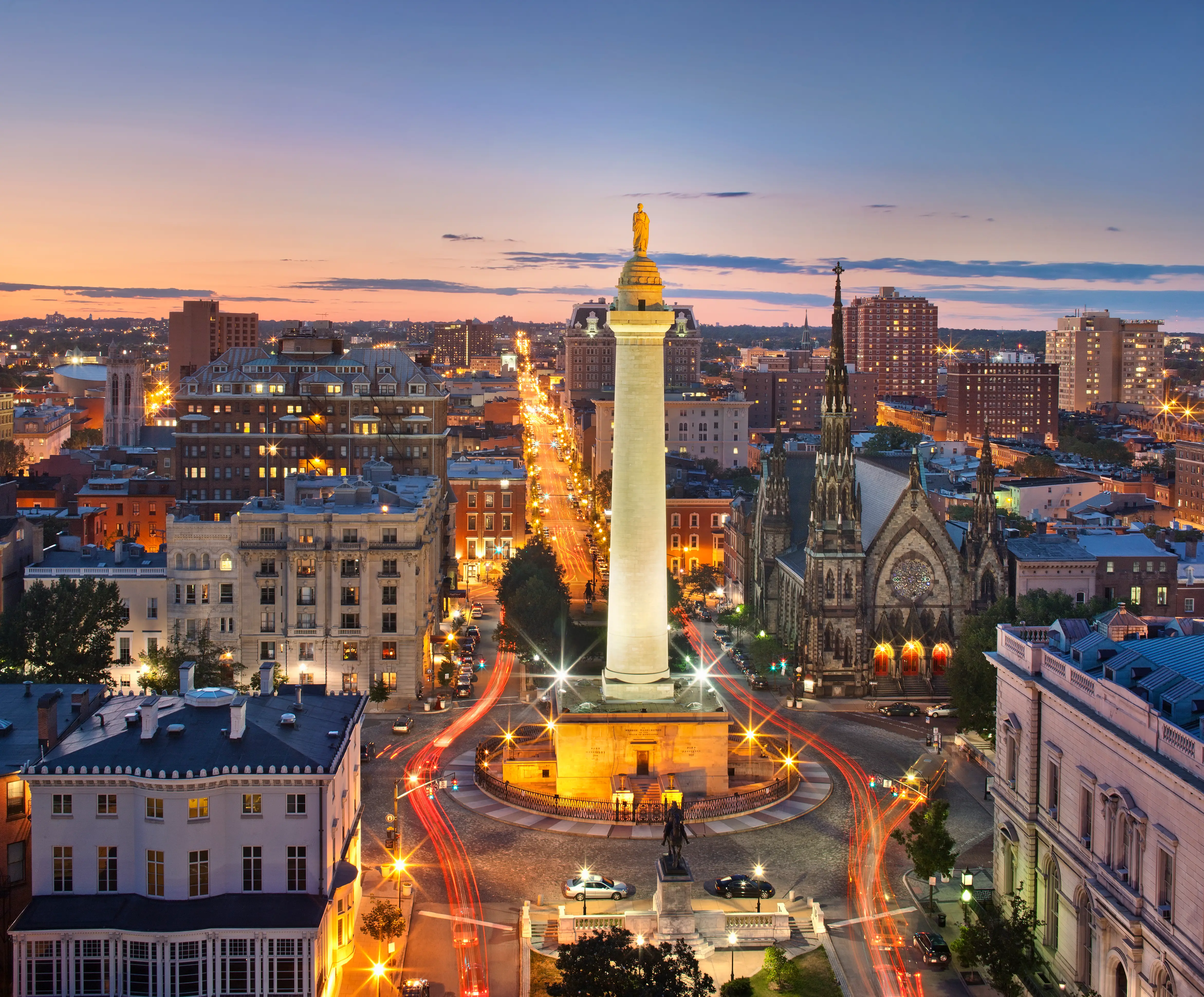 Baltimore’s Washington Monument rises over Mount Vernon streets and illuminated buildings at dusk.