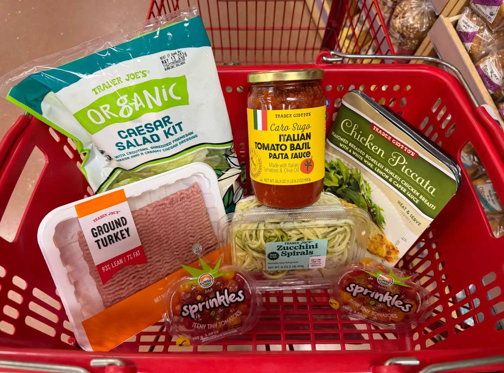 Trader Joe's groceries in a red shopping cart, including salad kit, ground turkey, pasta sauce, zucchini spirals, and tomatoes.