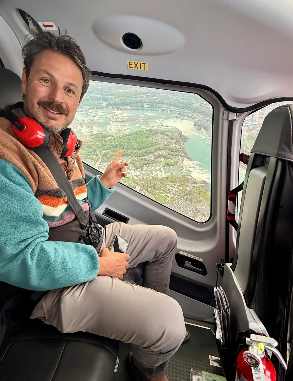 Man wearing a headset sits in a helicopter cabin and points toward an aerial river landscape through the window.