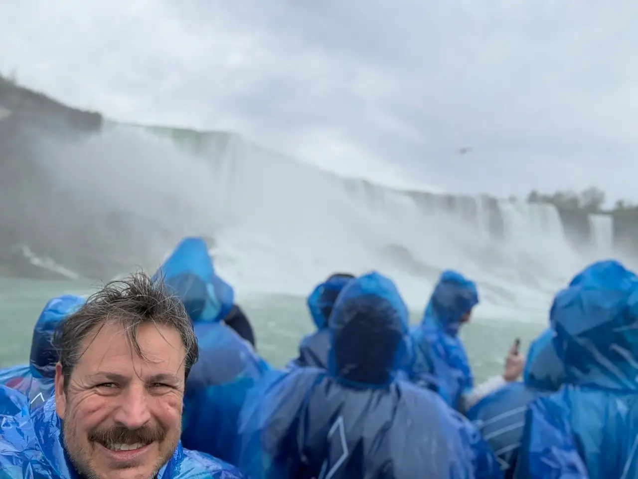 Smiling man in a blue poncho takes a selfie among tourists near the misty Niagara Falls.