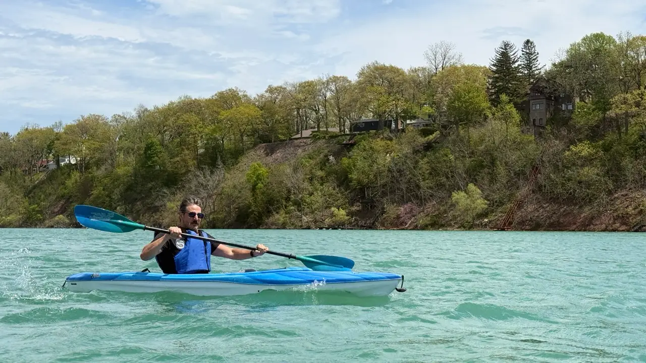 A person paddles a blue kayak across turquoise water below a wooded shoreline with houses on a bluff.