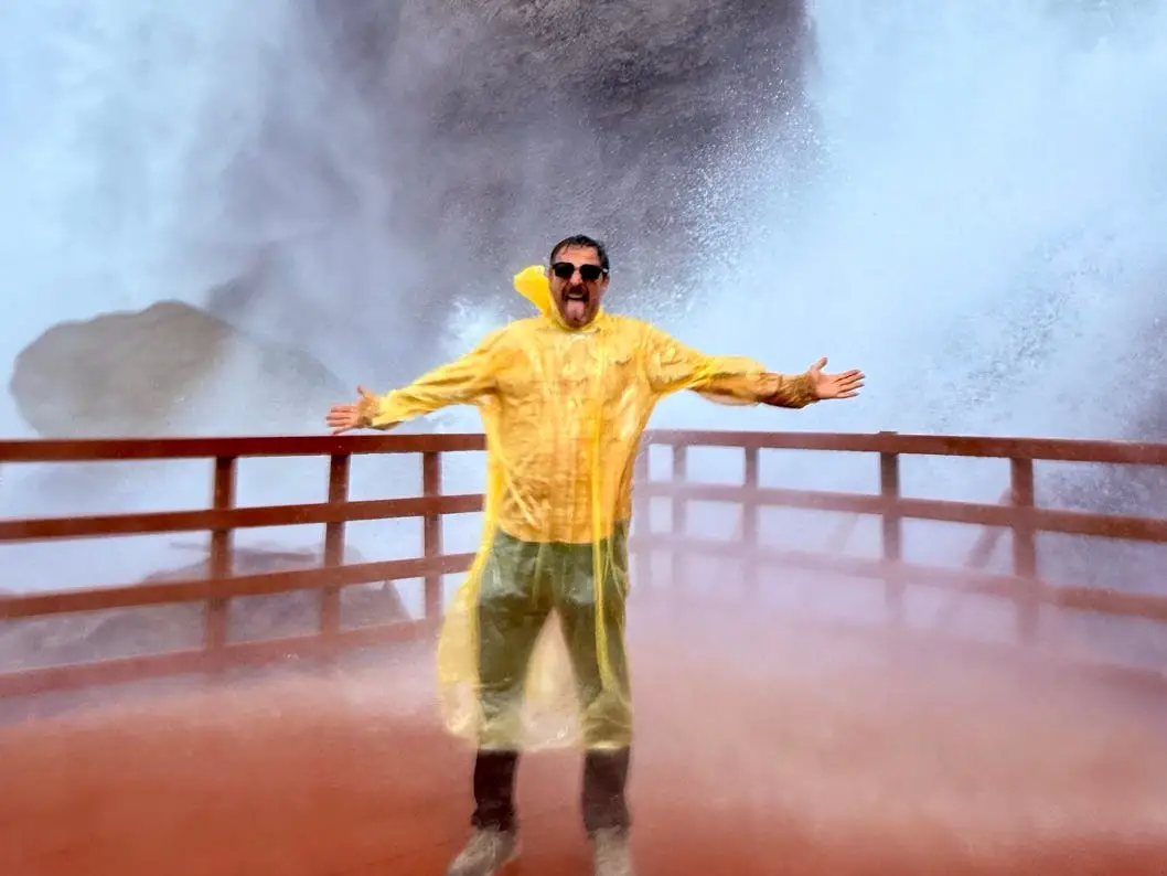 Person in a yellow rain poncho stands with arms out on a misty waterfall observation deck.
