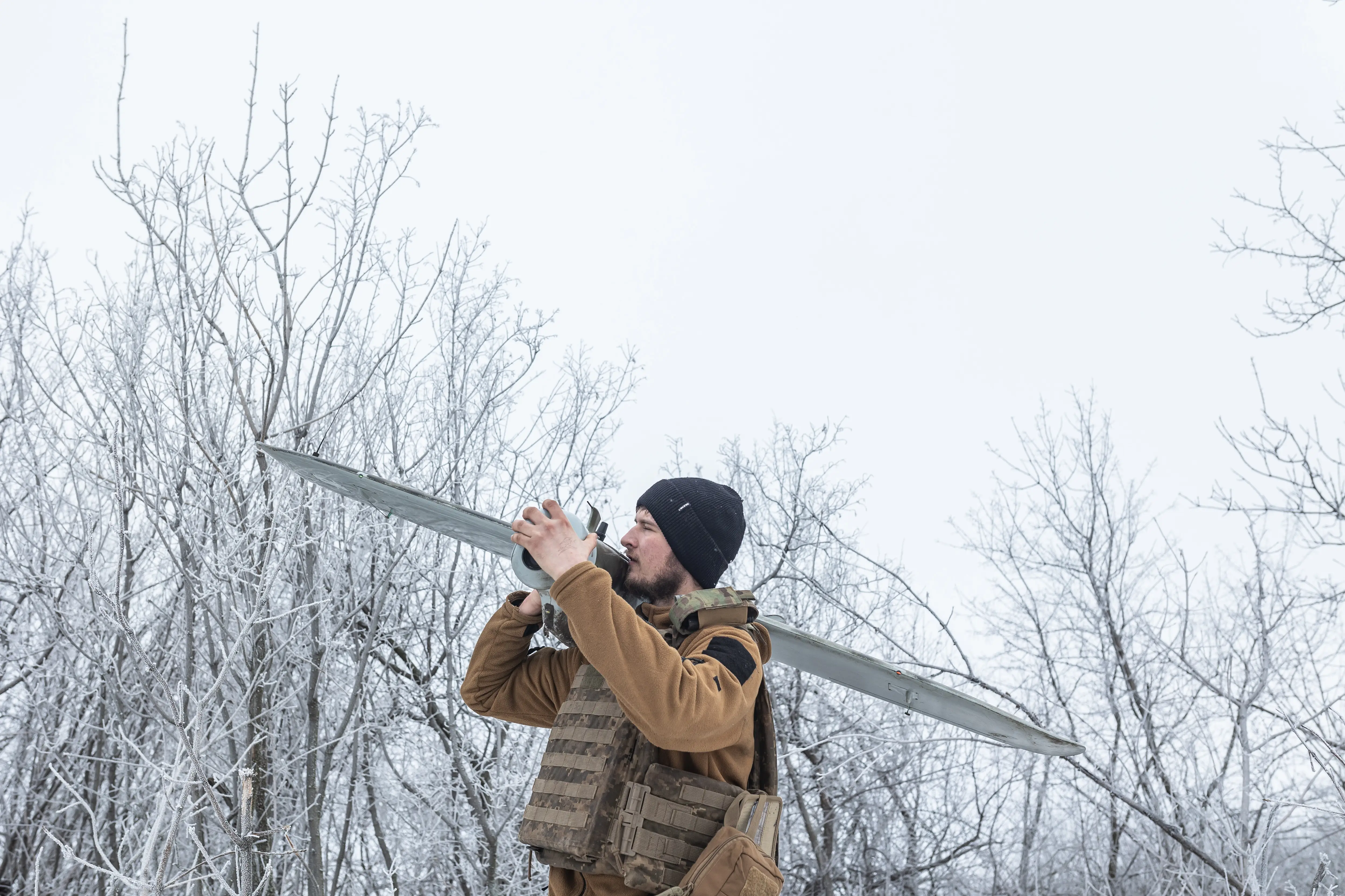 A man in khaki and a black beanie stands in a snowy field with trees with a large grey drone over his shoulder