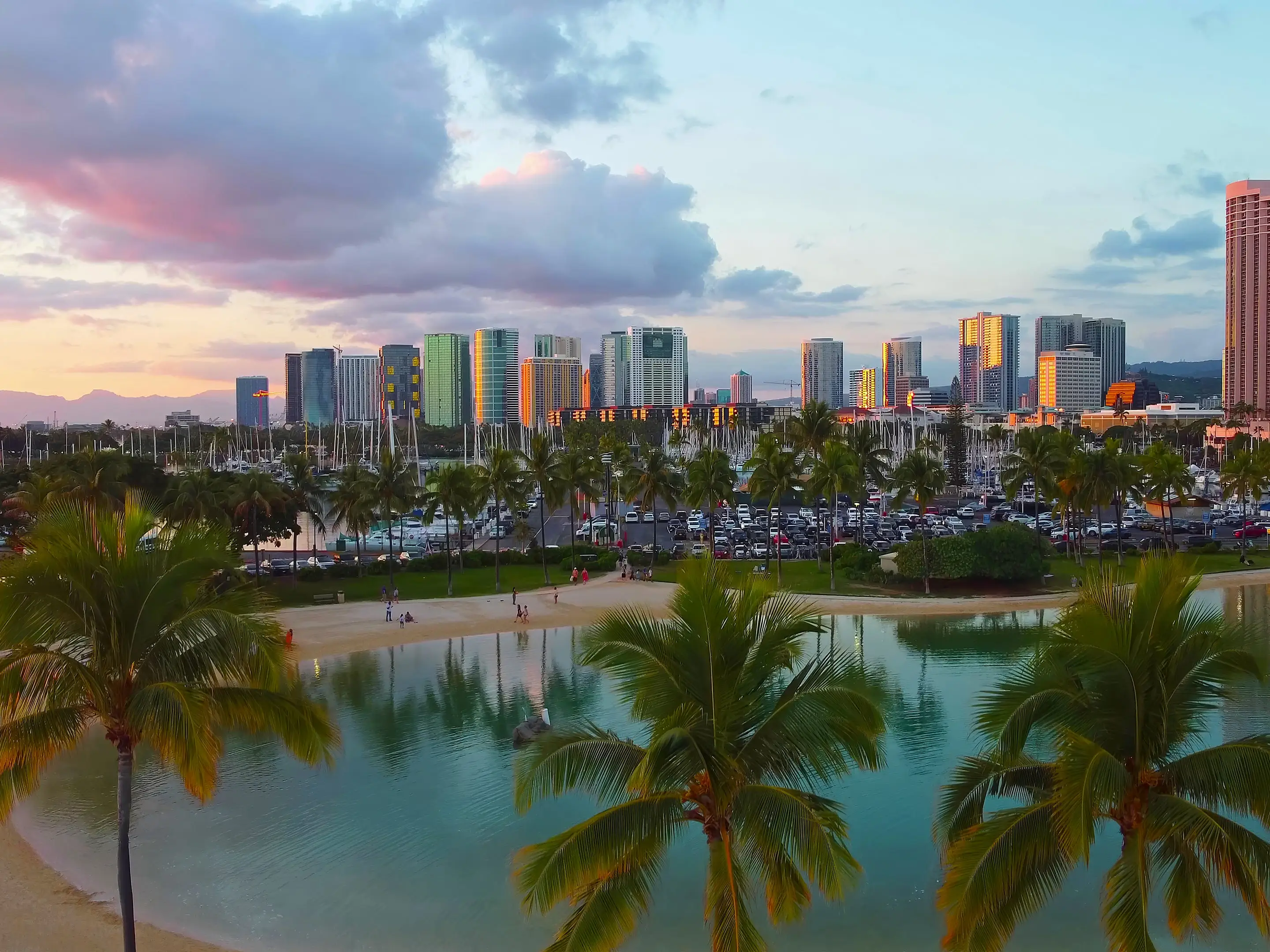 Honolulu skyline and lagoon at sunset. Scenic view of a tropical lagoon with palm trees, sandy beach, and the illuminated Honolulu skyline reflecting the warm hues of sunset