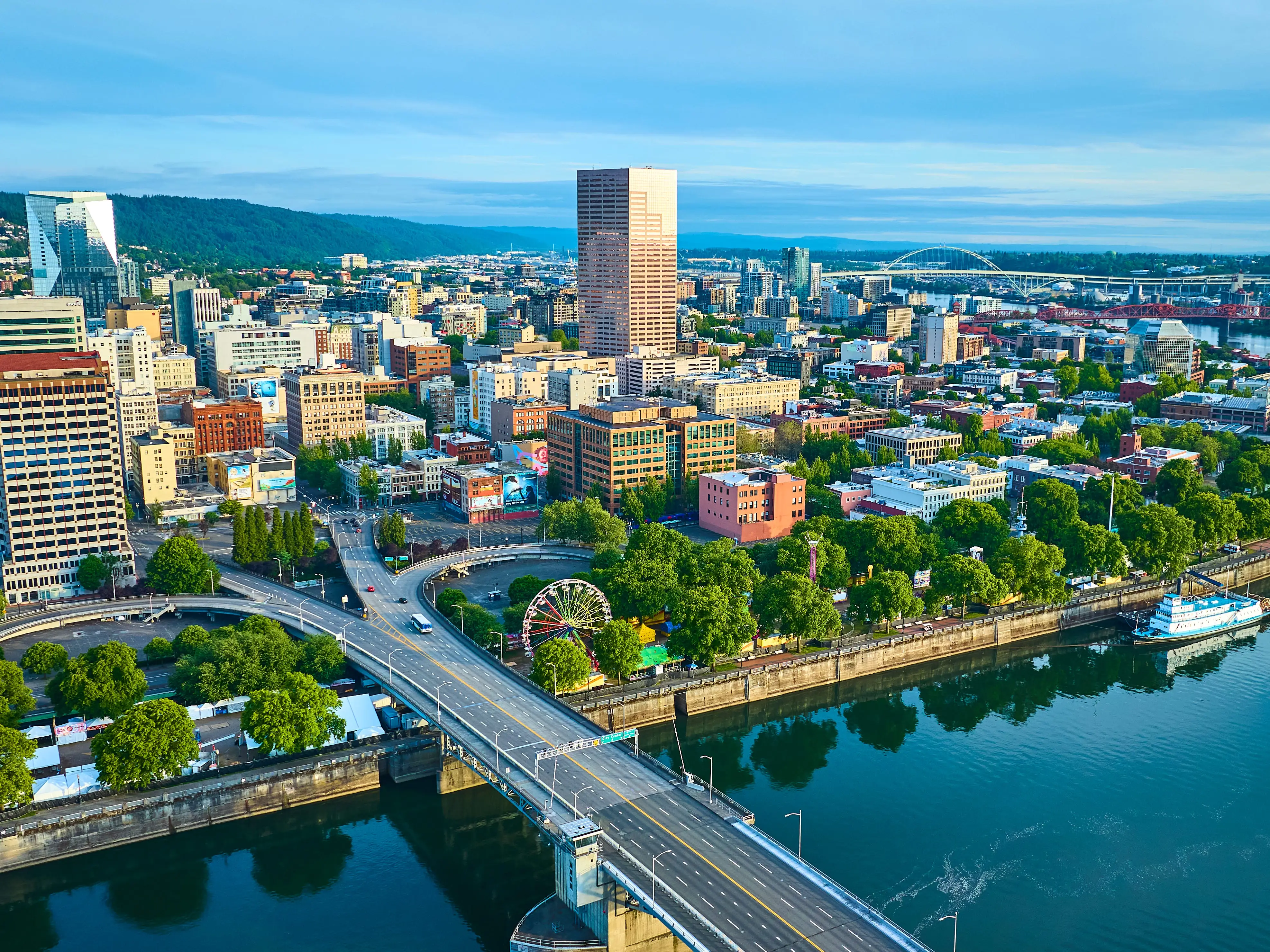 Aerial view of Portland, Oregon, showcasing the vibrant downtown with a mix of modern and classic skyscrapers. The Willamette River with a bridge and paddlewheel riverboat adds scenic beauty and historical charm.