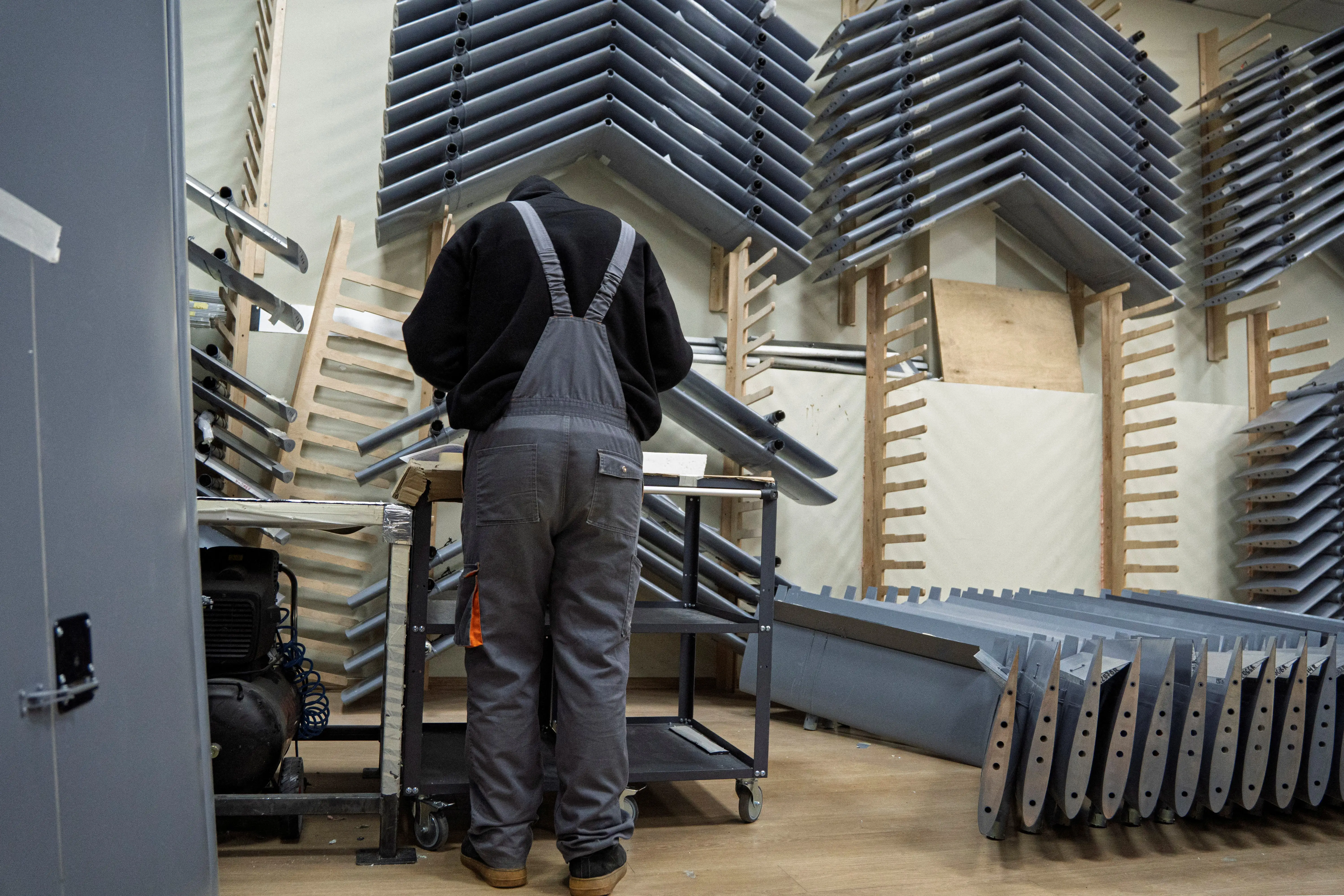 A man in grey dungarees and a black top stands with his back turned looking at a wall of large grey drone wings
