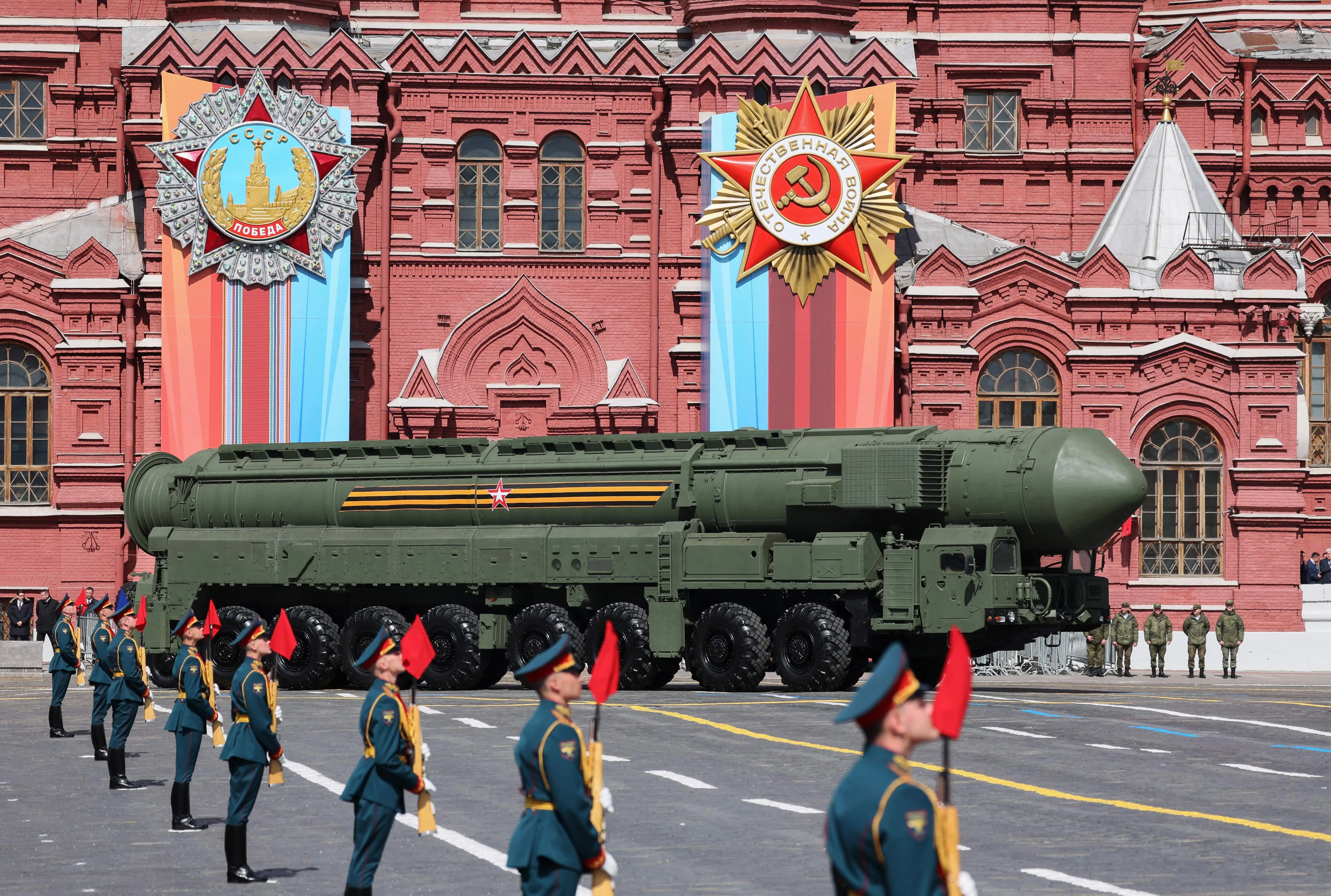 A large green missile system on wheels on a road in front of an ornate red building with people in military dress standing in a line