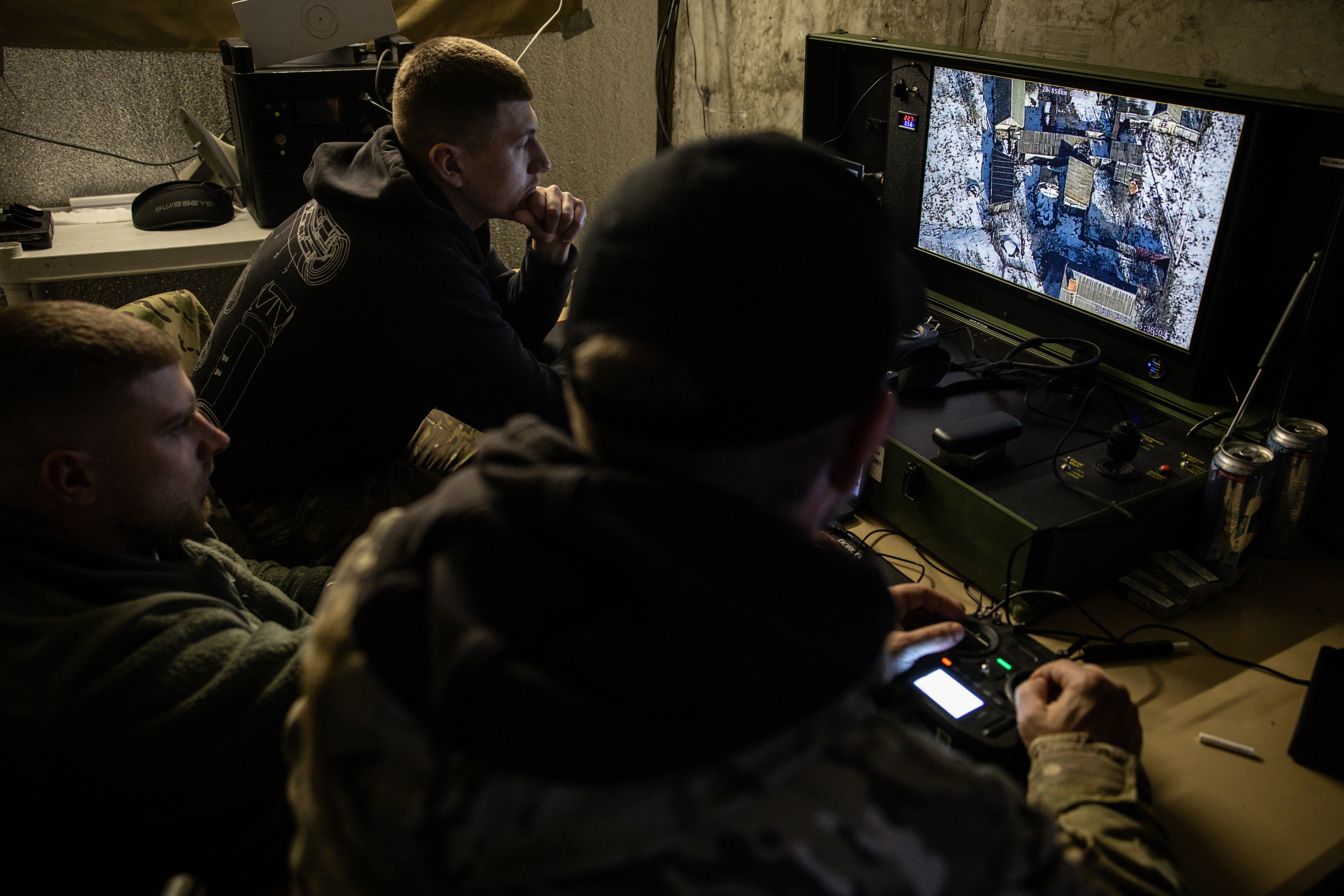 Three men in camouflage gear in a small, dark indoor location look at a screen that has aerial footage on it