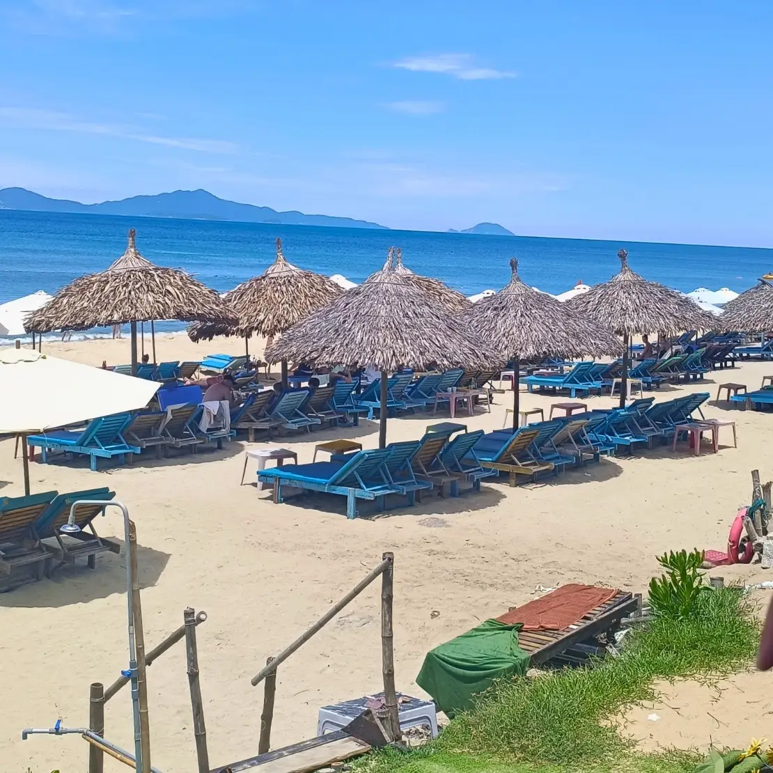 Sandy beach lined with thatched umbrellas and blue lounge chairs beside calm ocean water and distant islands.