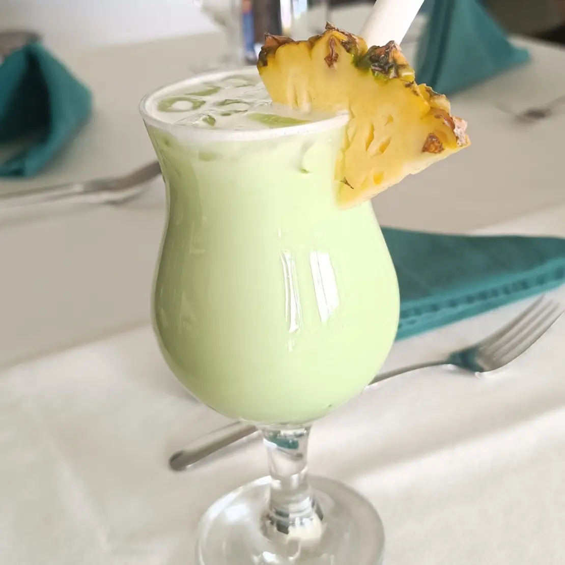 Pale green tropical drink in a stemmed glass garnished with pineapple on a restaurant table.