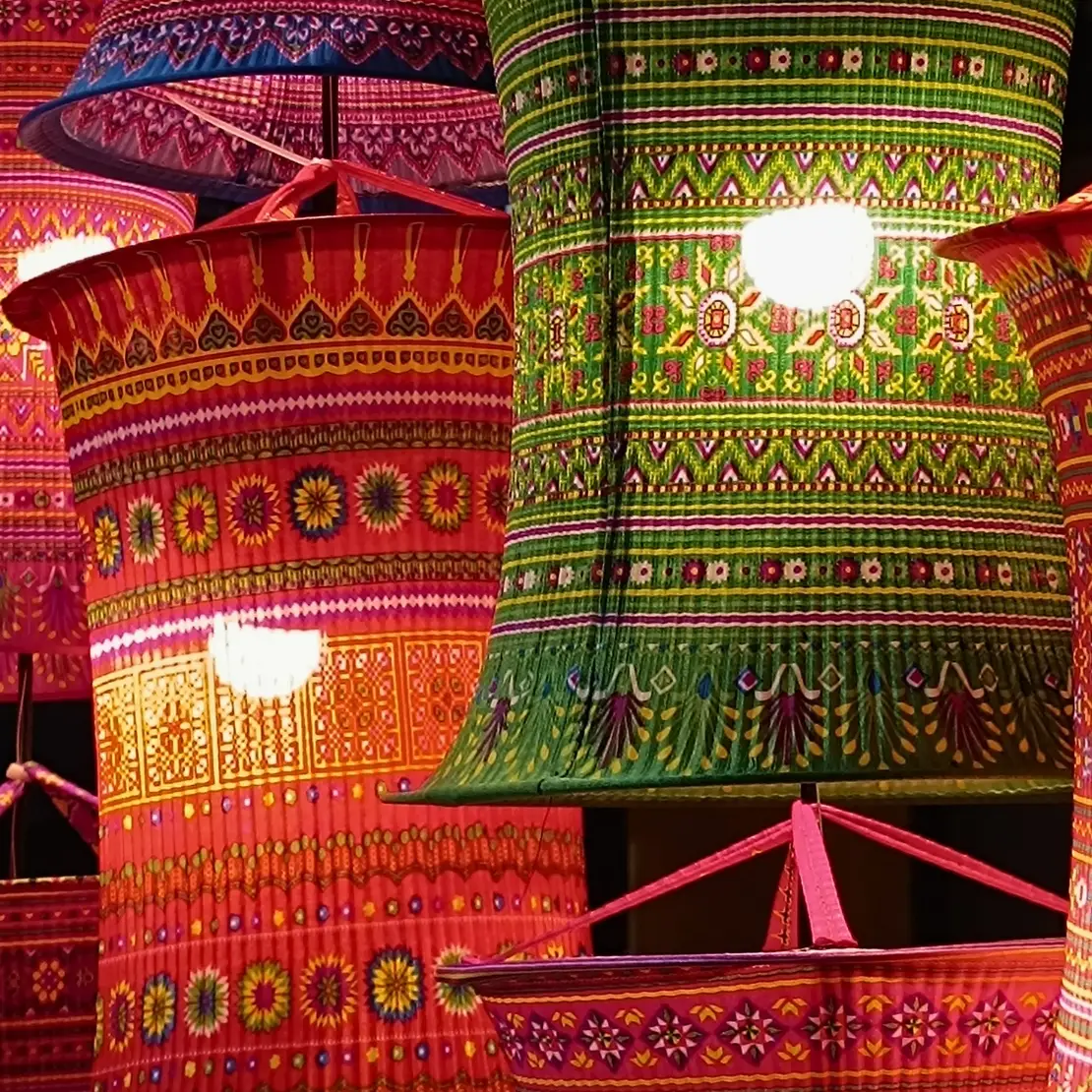 Colorful patterned fabric lanterns hang closely together, glowing with warm light in red, green, pink, and orange.