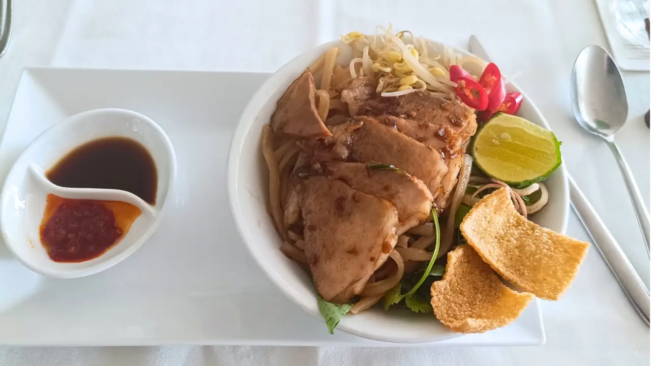 Bowl of noodles with sliced chicken, bean sprouts, lime, chilies, crackers, and sauces on a white table setting.