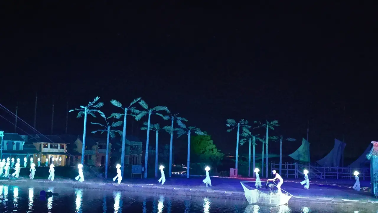 Illuminated performers move along a waterfront stage at night with palm trees, reflections, and a boat prop.