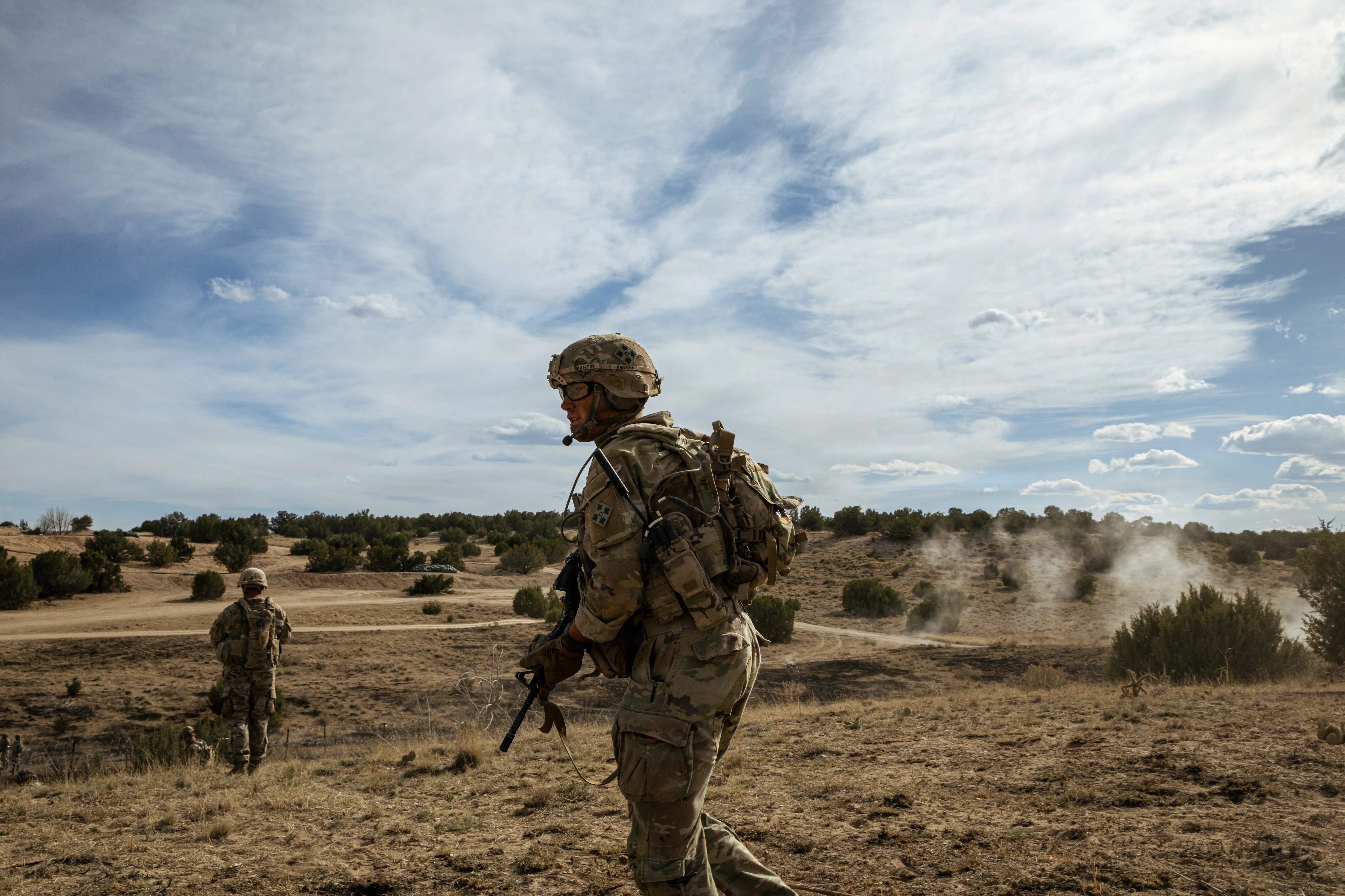 Soldiers wearing camouflage walk along a dry field with a cloudy blue sky in the background.