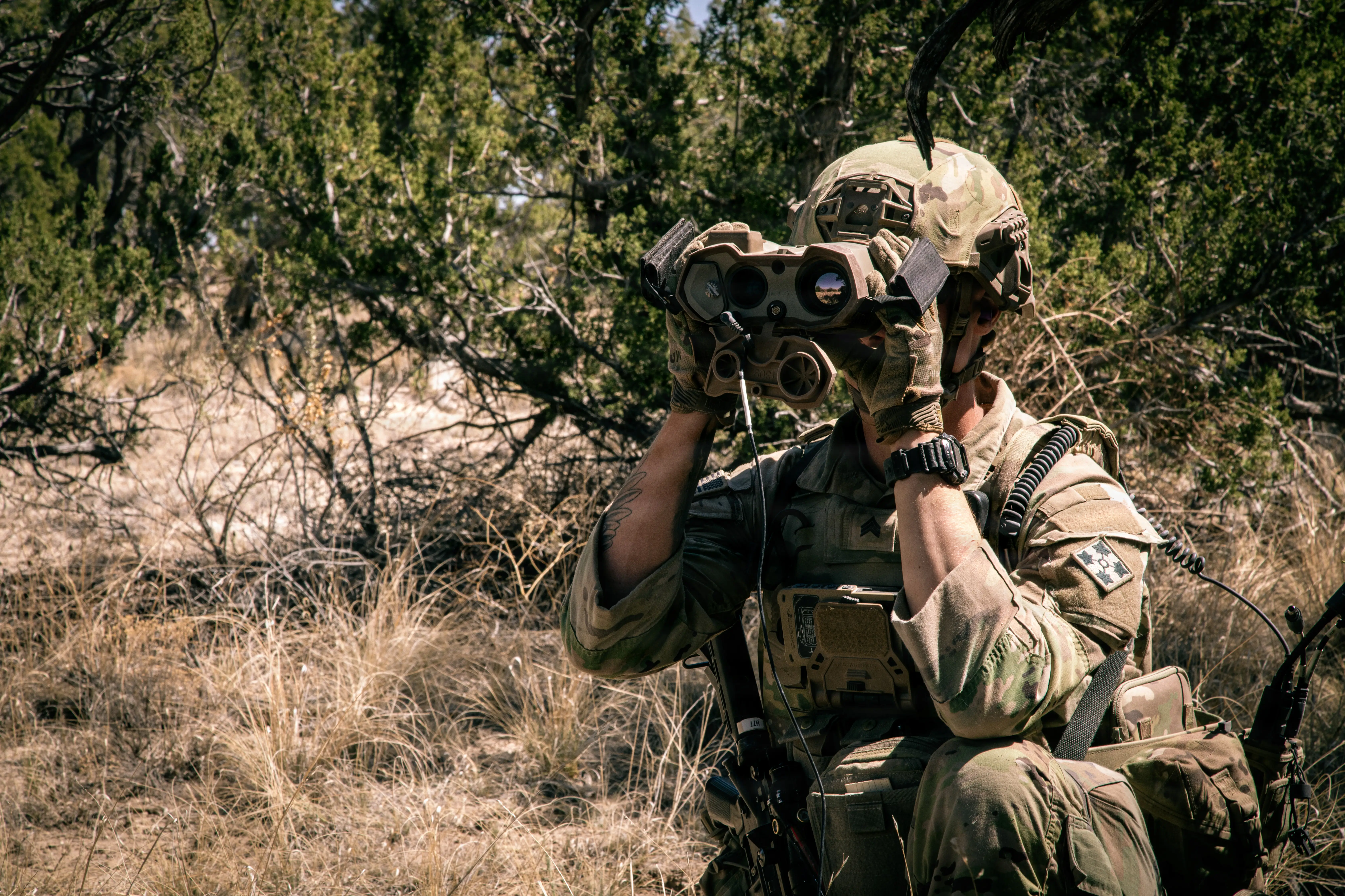 A soldier wearing camouflage looks through binoculars. Behind him is a line of green trees.
