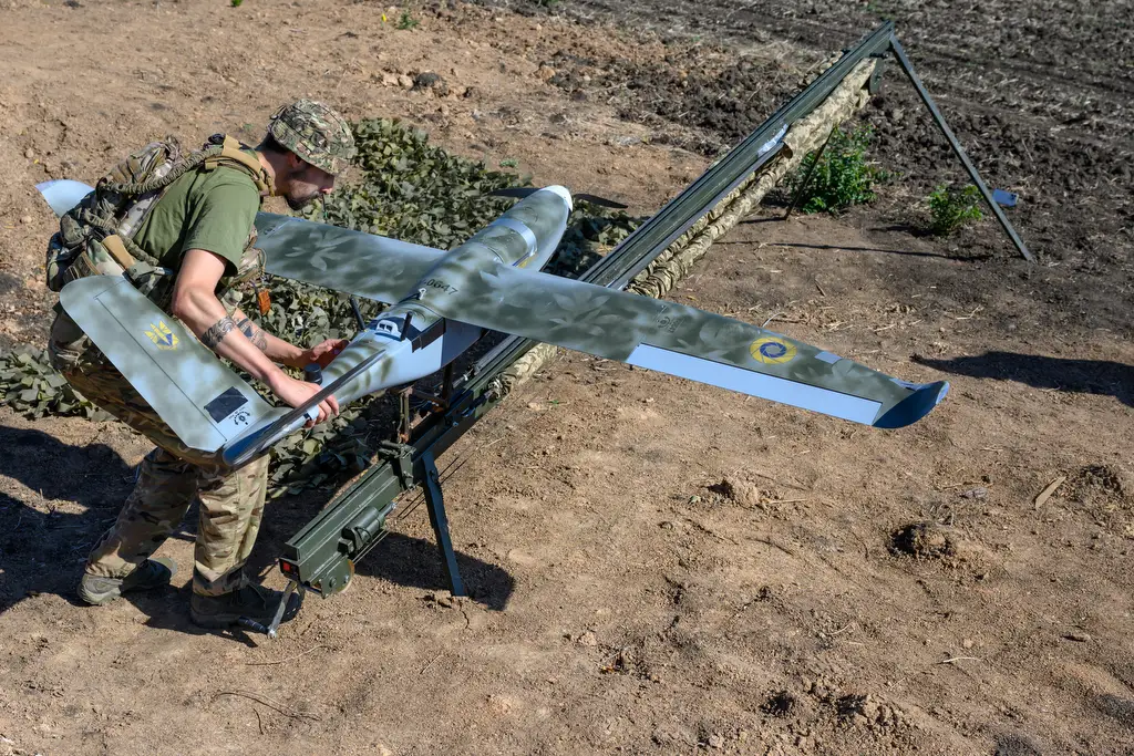 A man in khaki holds a drone painted with camouflage over a brown ground