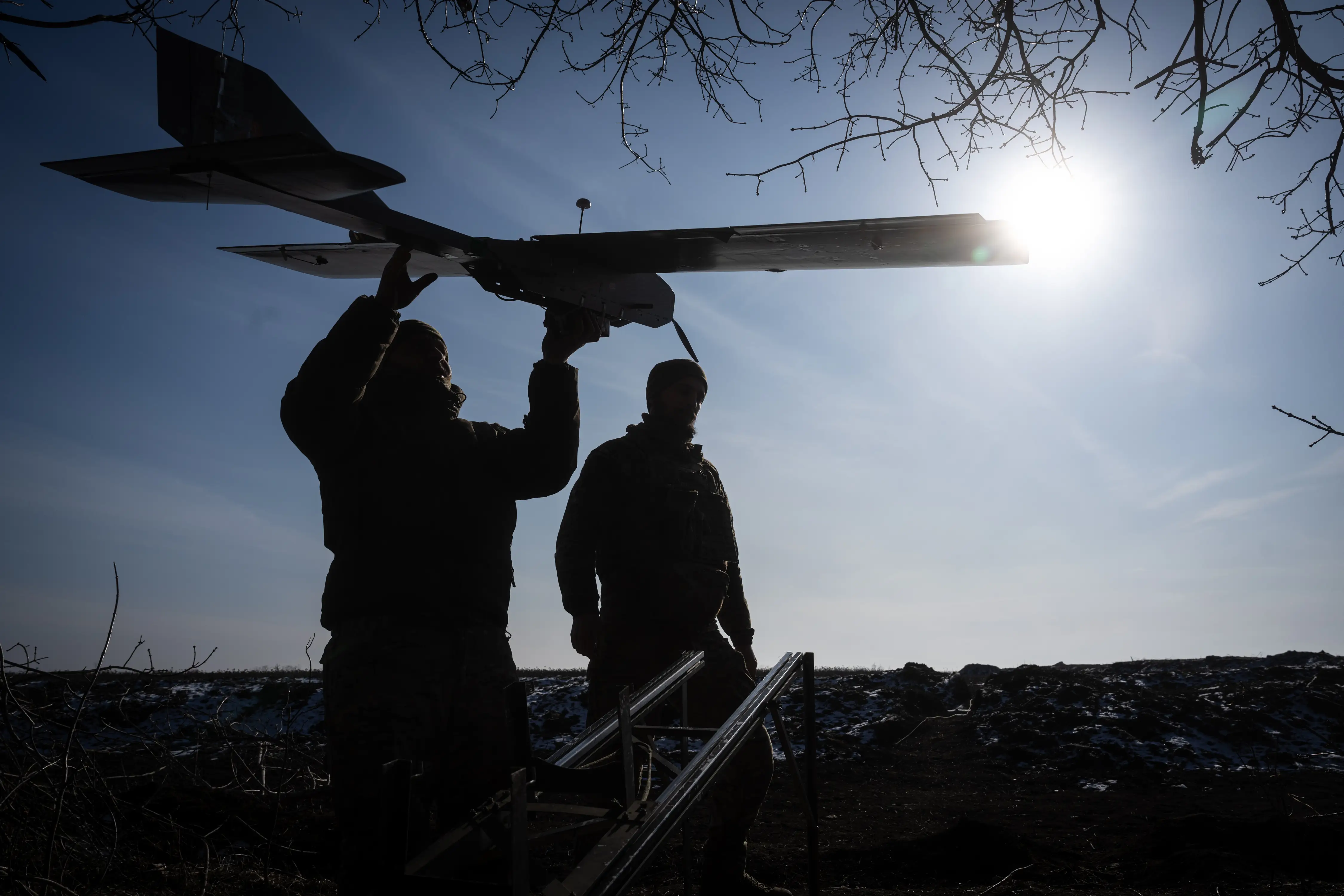 A man holds a drone against a sunny sky, with another man beside him. Both are silhouetted