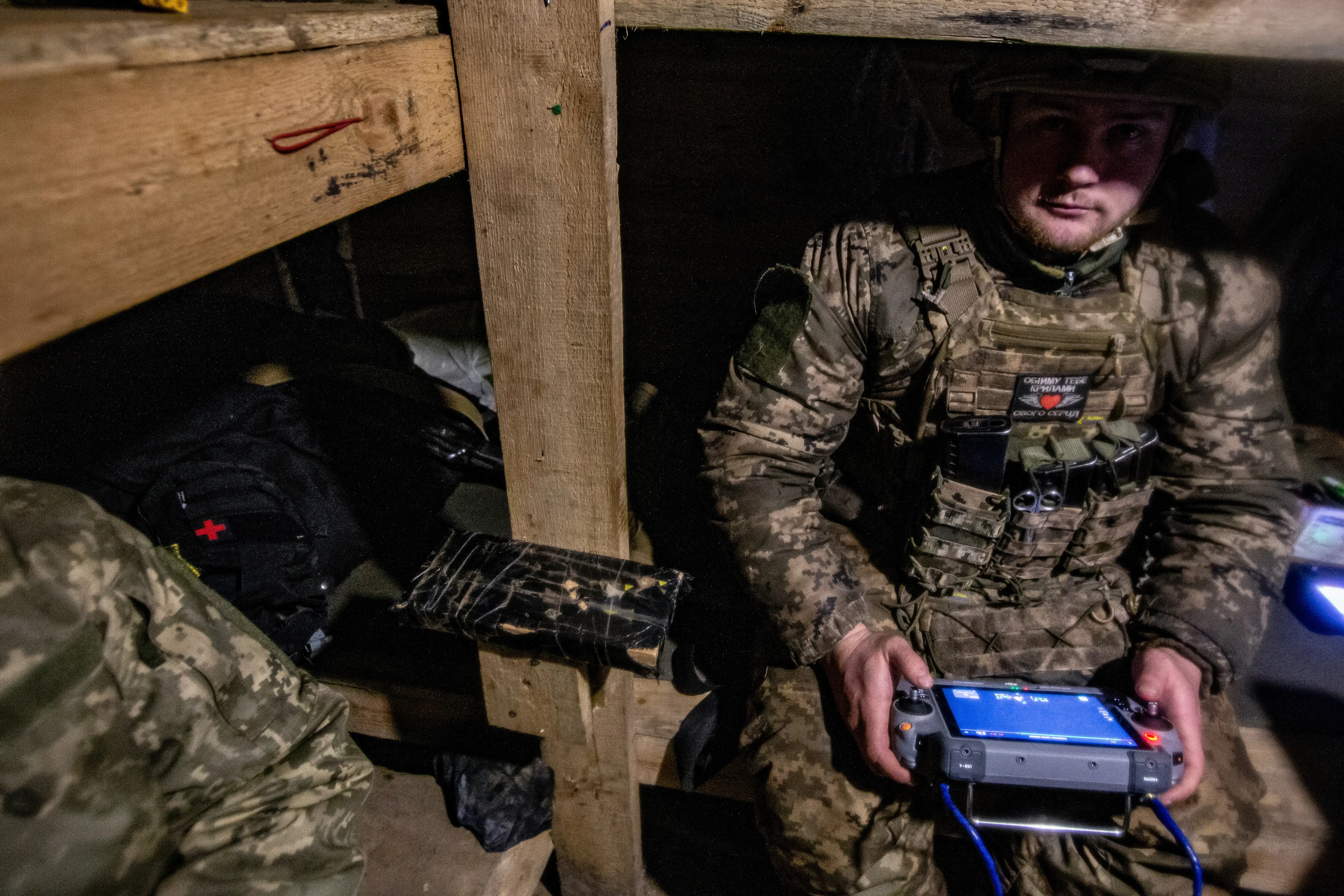 A man in camouflage gear sits on a wooden structure holding a controller in a small indoor space
