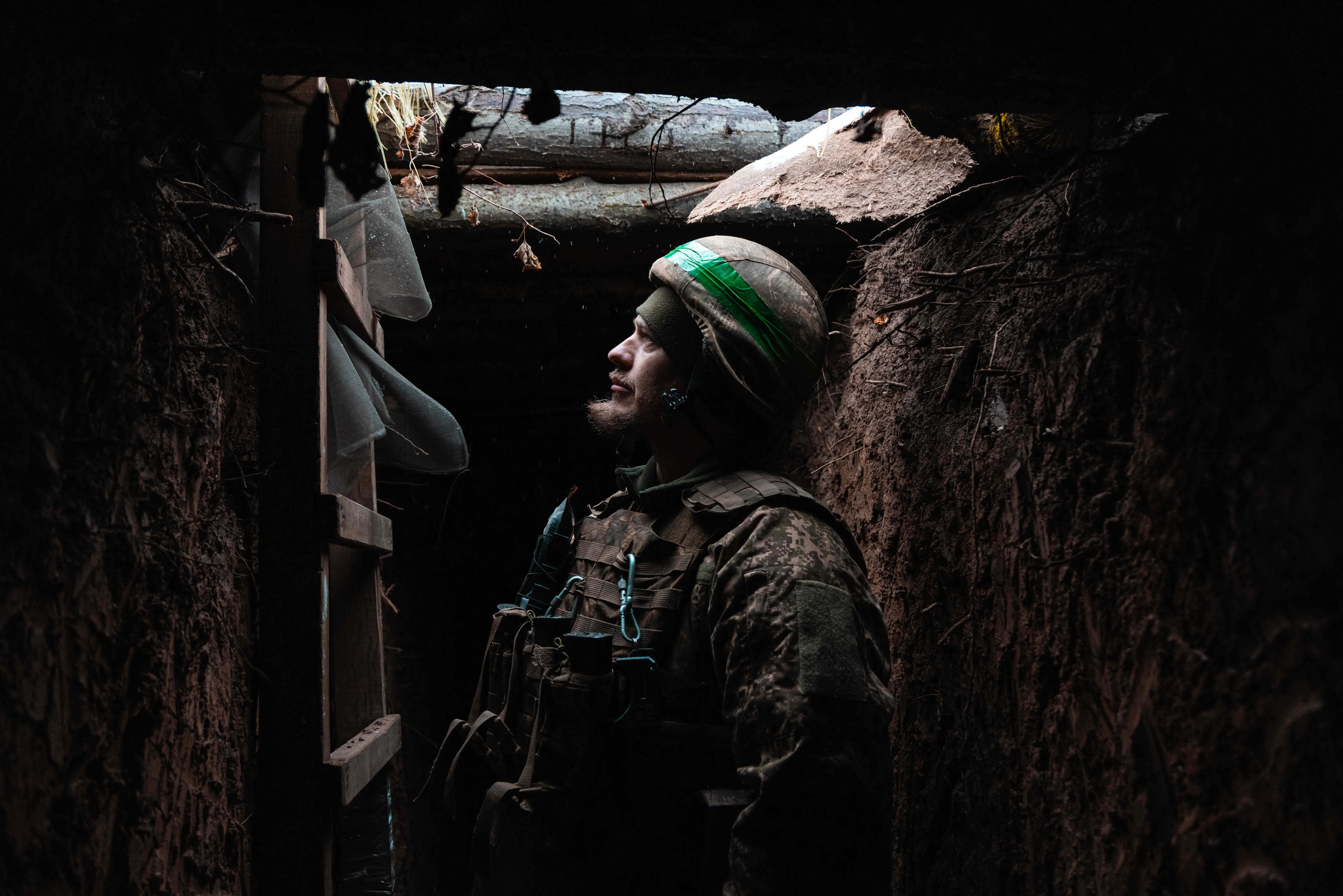A man in camouflage gear and a helmet and with a beard stands in a trench with a wooden ladder in front of him