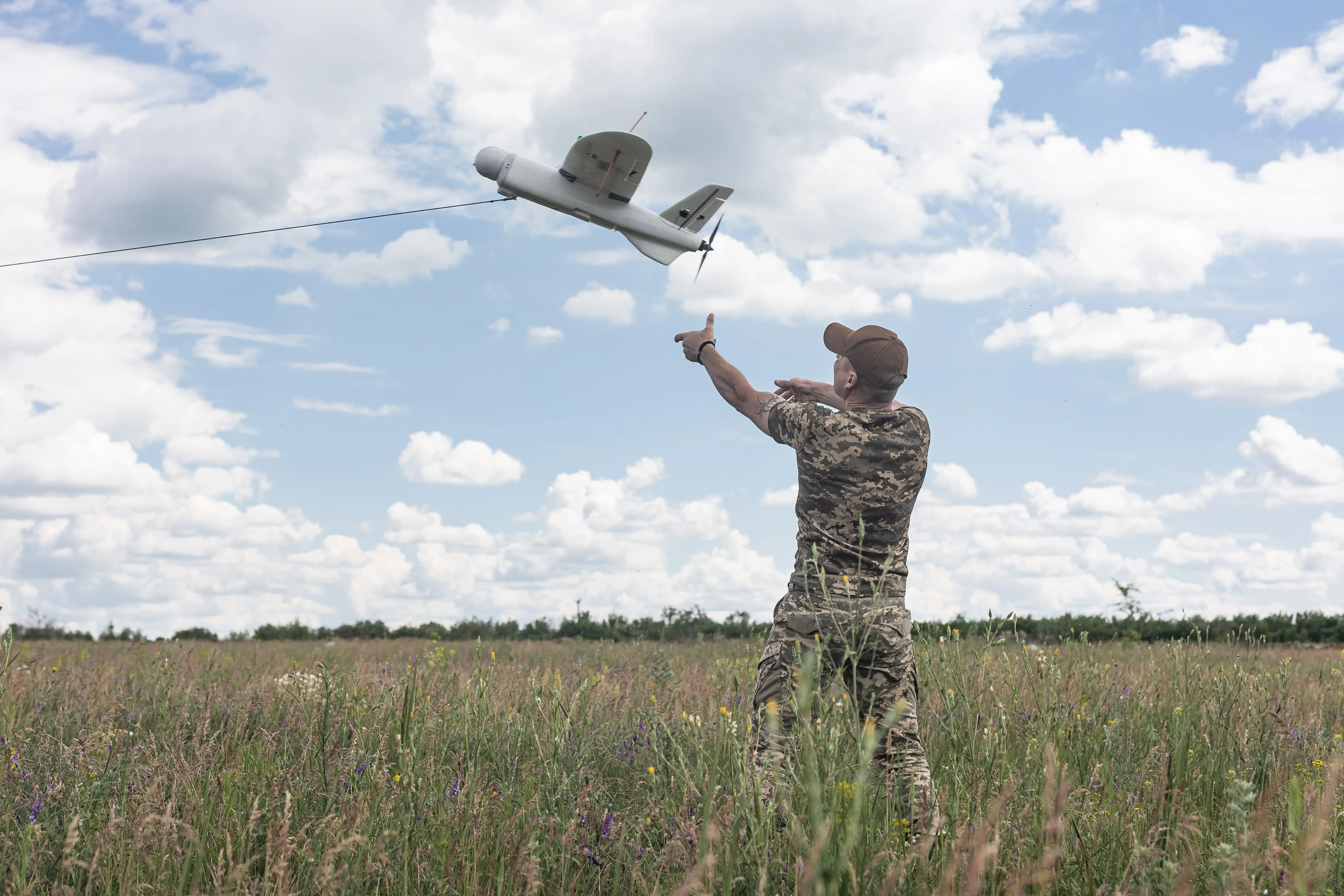A man in a camouflage t-shirt and trousers and wearing a cap stands in a field with his arms outstretched to a grey drone i the air under a blue and cloudy sky.