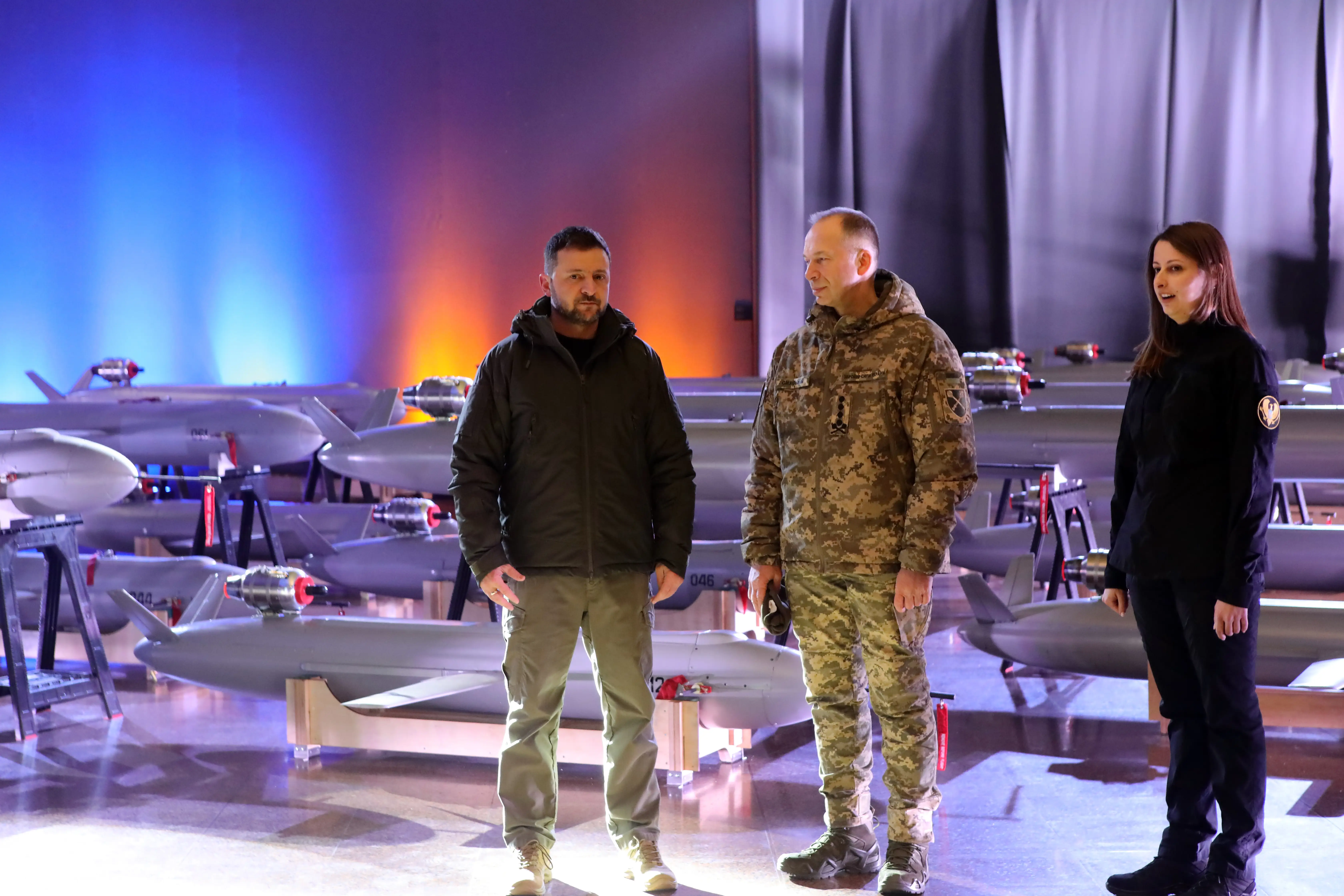 Three figures stand in a room surrounded by grey drones