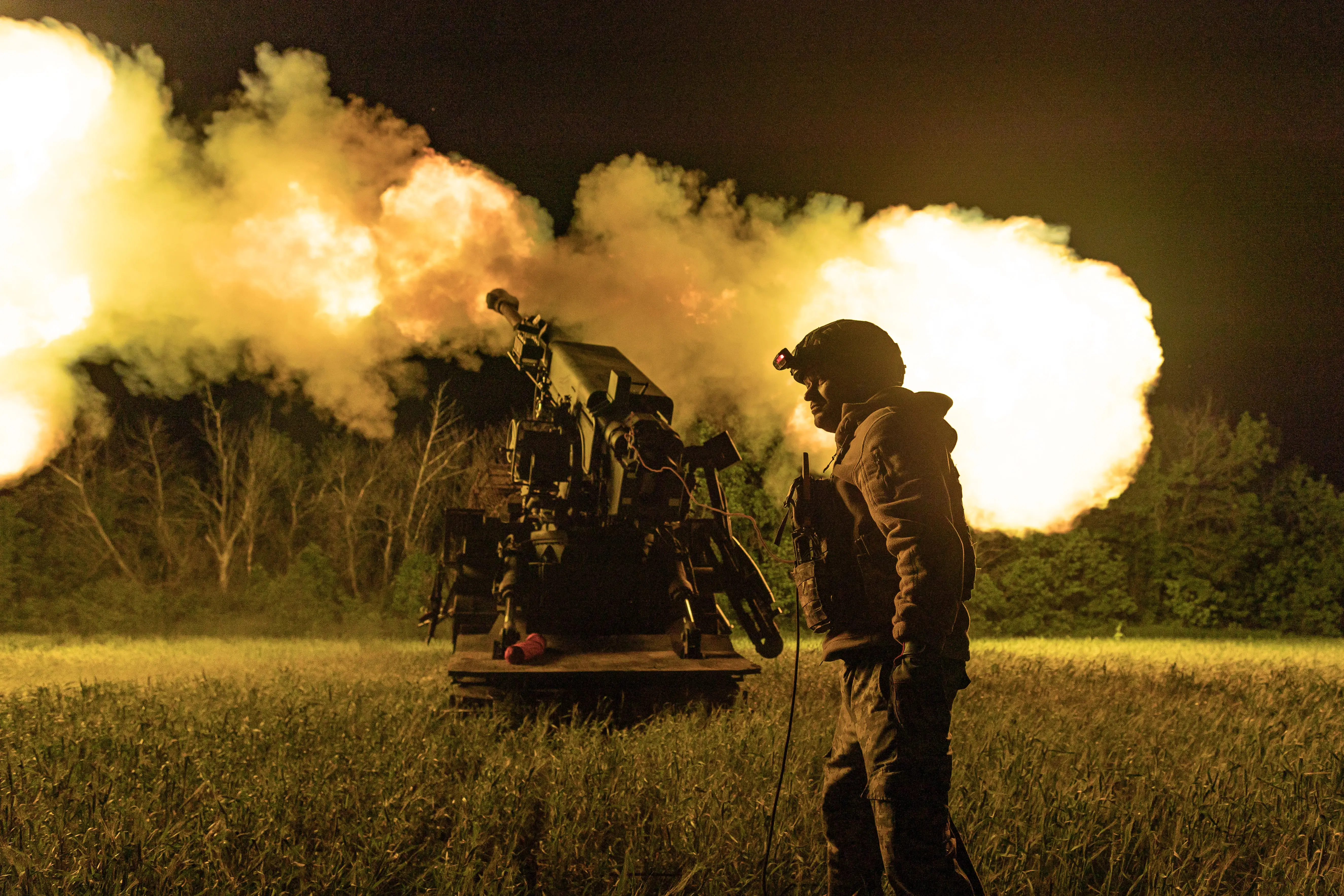 A man wearing khaki and a helmet stands in a field at night as a large piece of weaponry fires in front of him, creating clouds of orange smoke