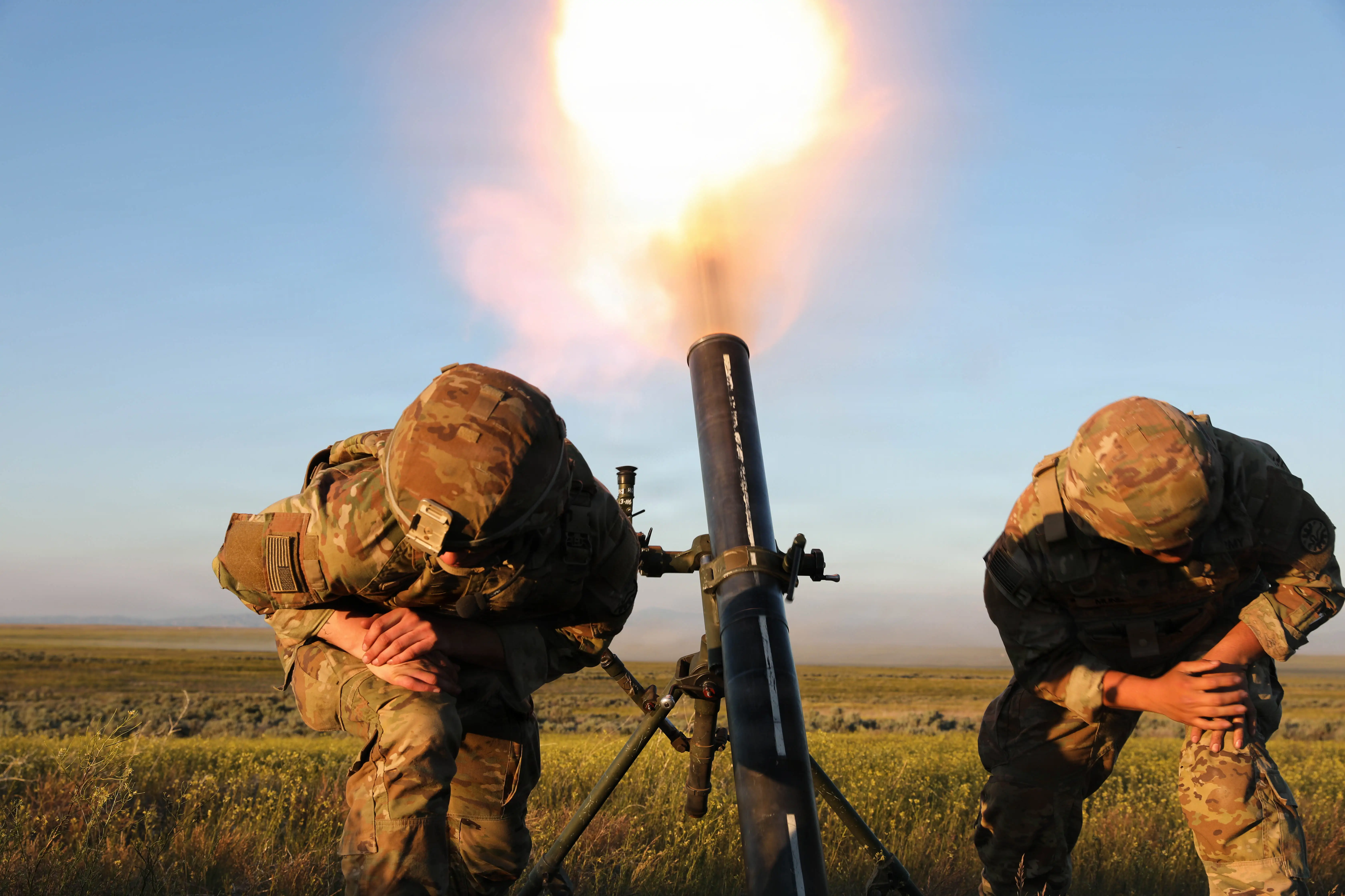 Troops wearing camouflage crouch as they fire a mortar into a clear blue sky.