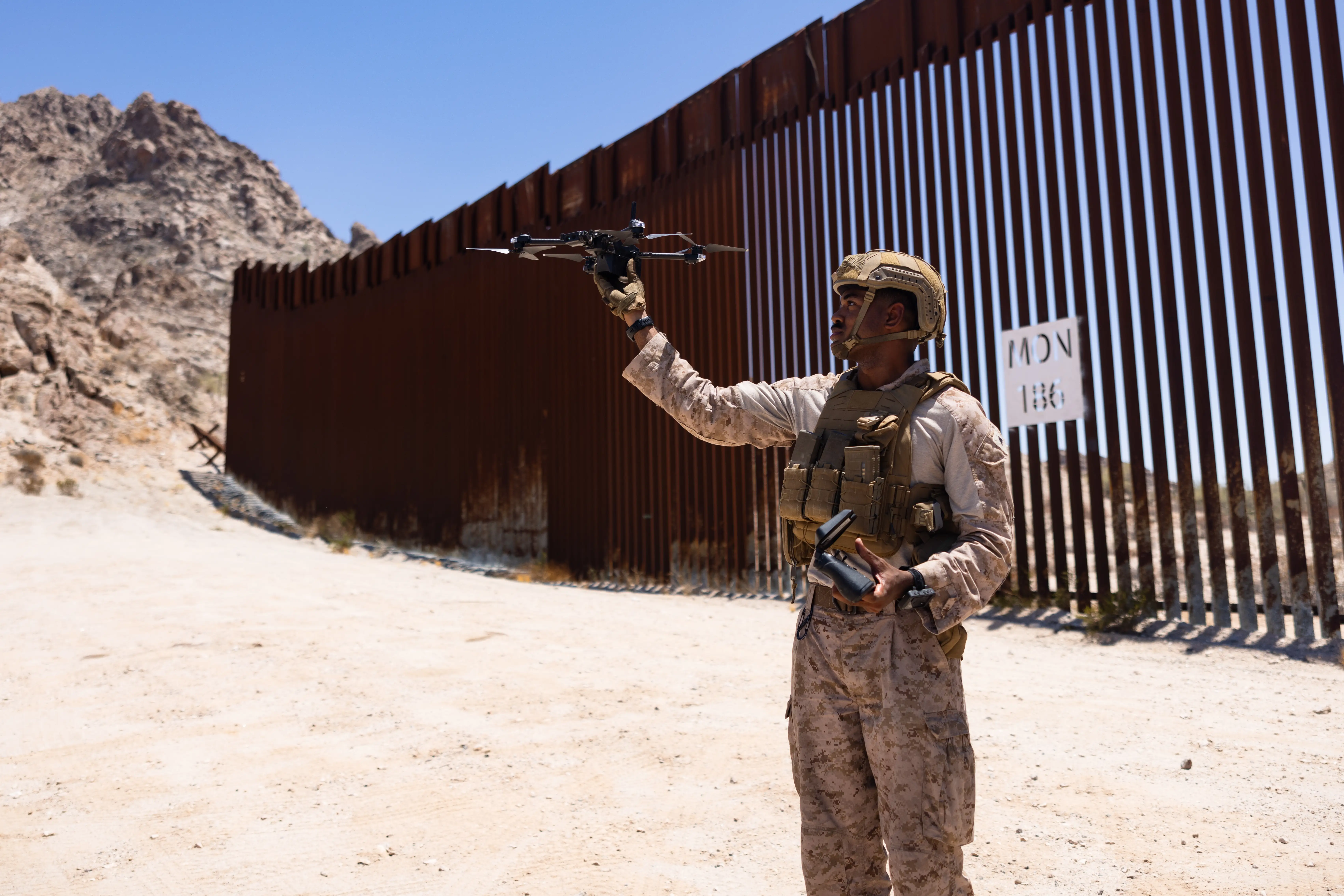 A Marine wearing camouflage holds a small drone up while standing near the US southern boarder.