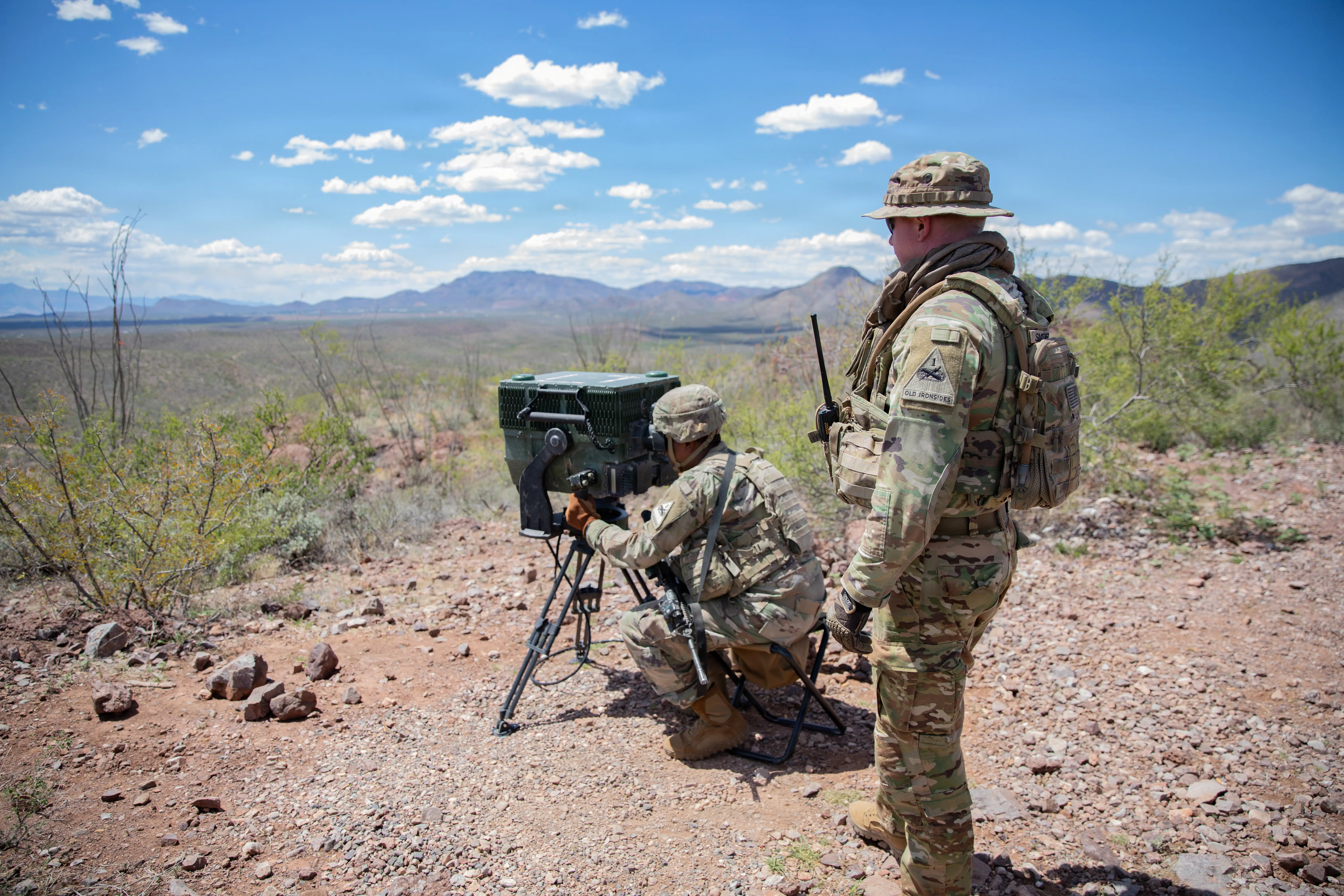 Two soldiers wearing camouflage are positioned on a hill in the desert. One is crouched and looking into a large, standing system.