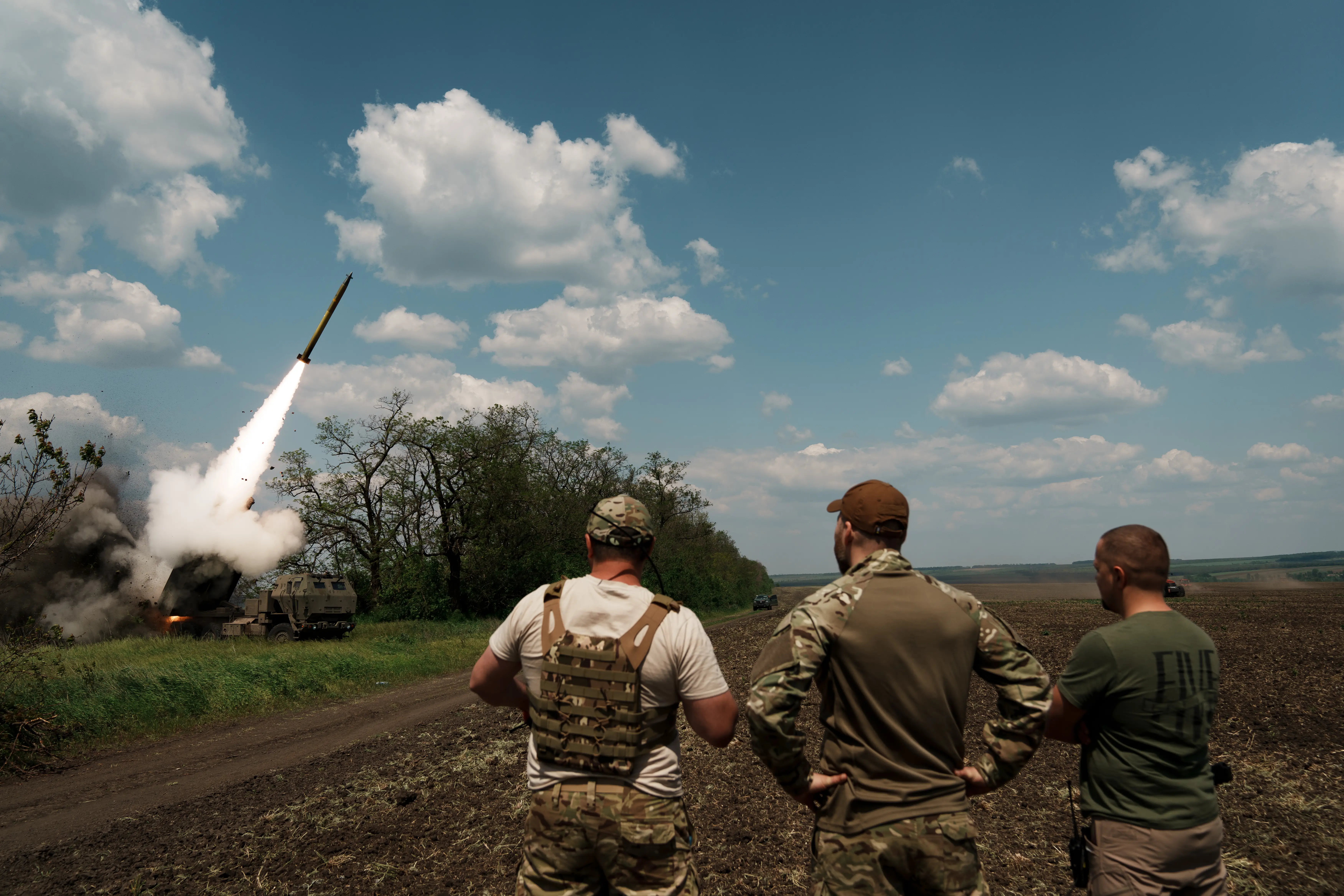 Three men look on as a HIMARS system fires its rocket.
