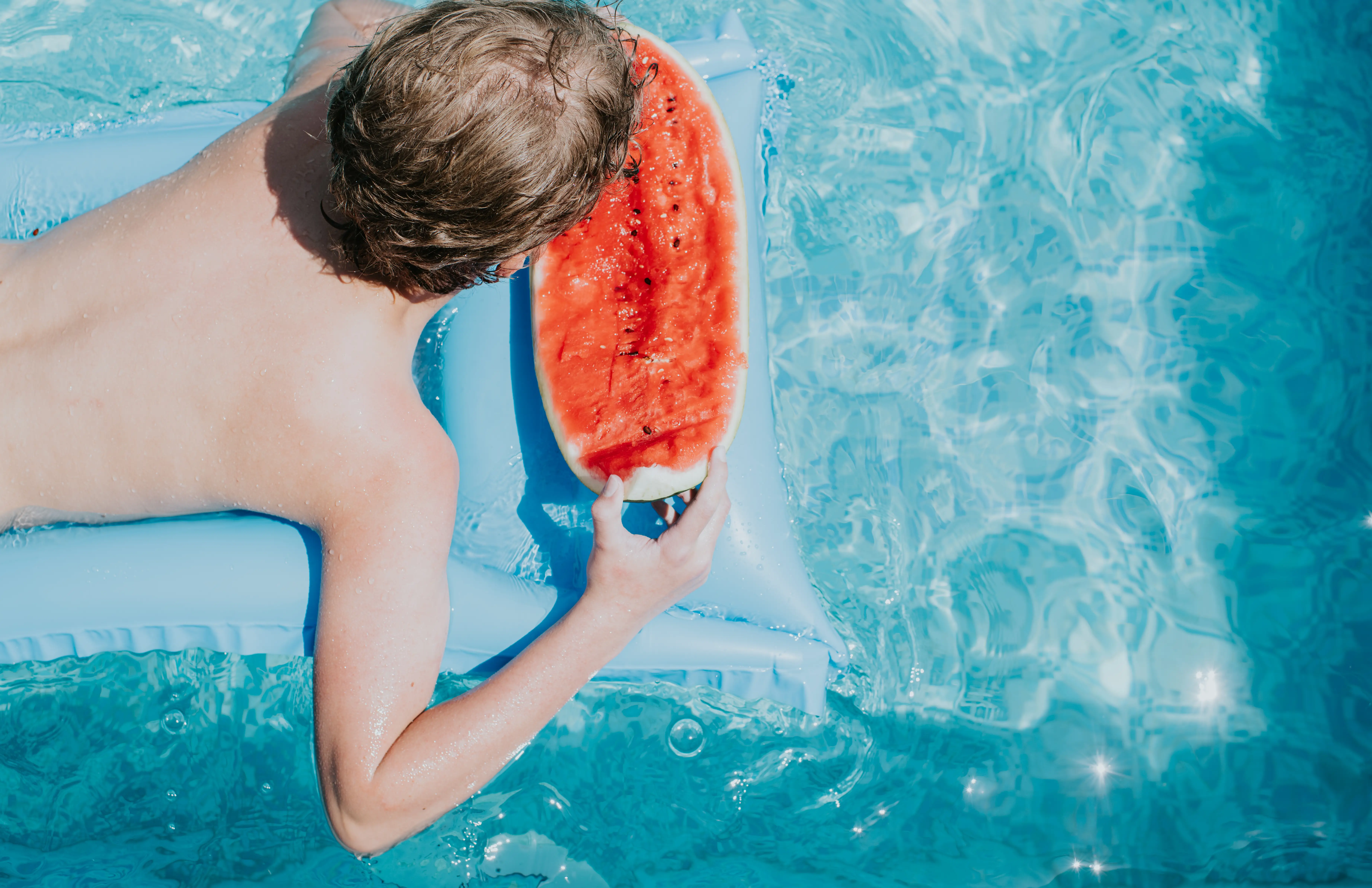 A boy snacks on watermelon while floating in a pool.