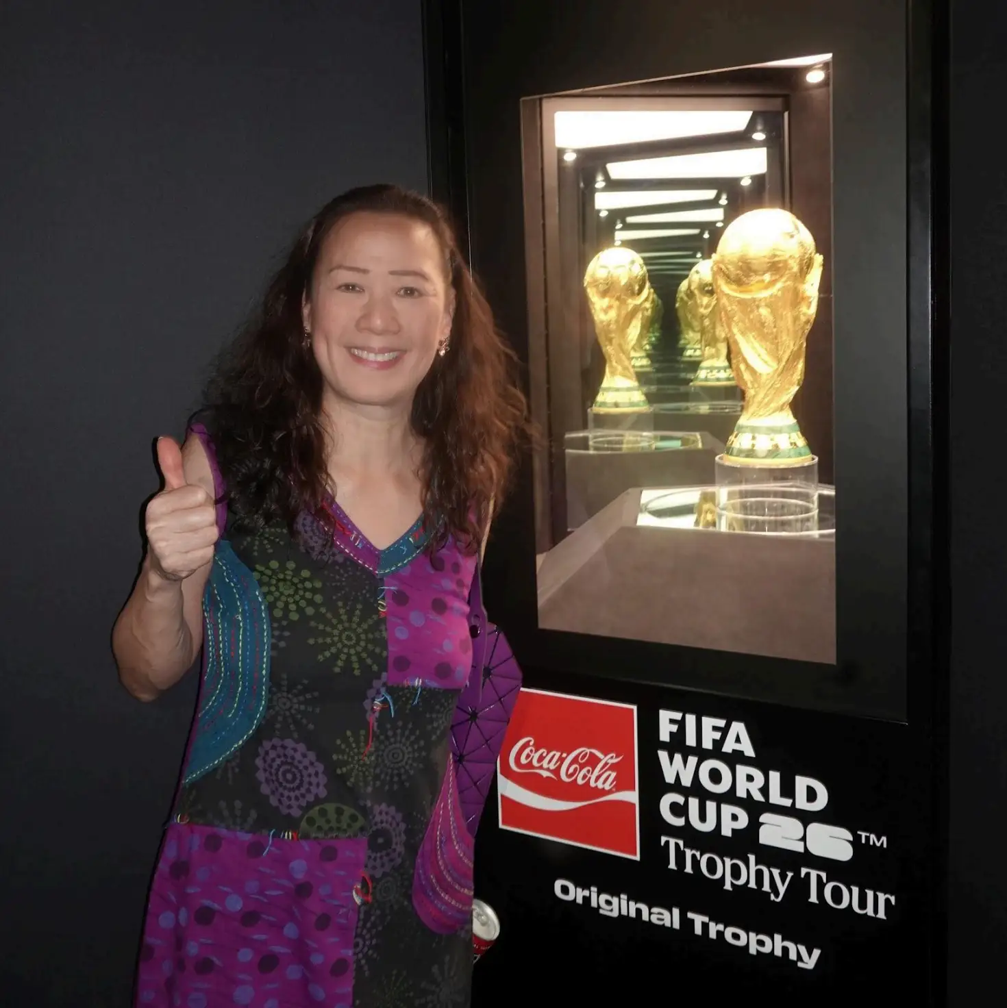 May Lee giving a thumbs-up beside a displayed FIFA World Cup trophy in a Coca-Cola branded tour case.