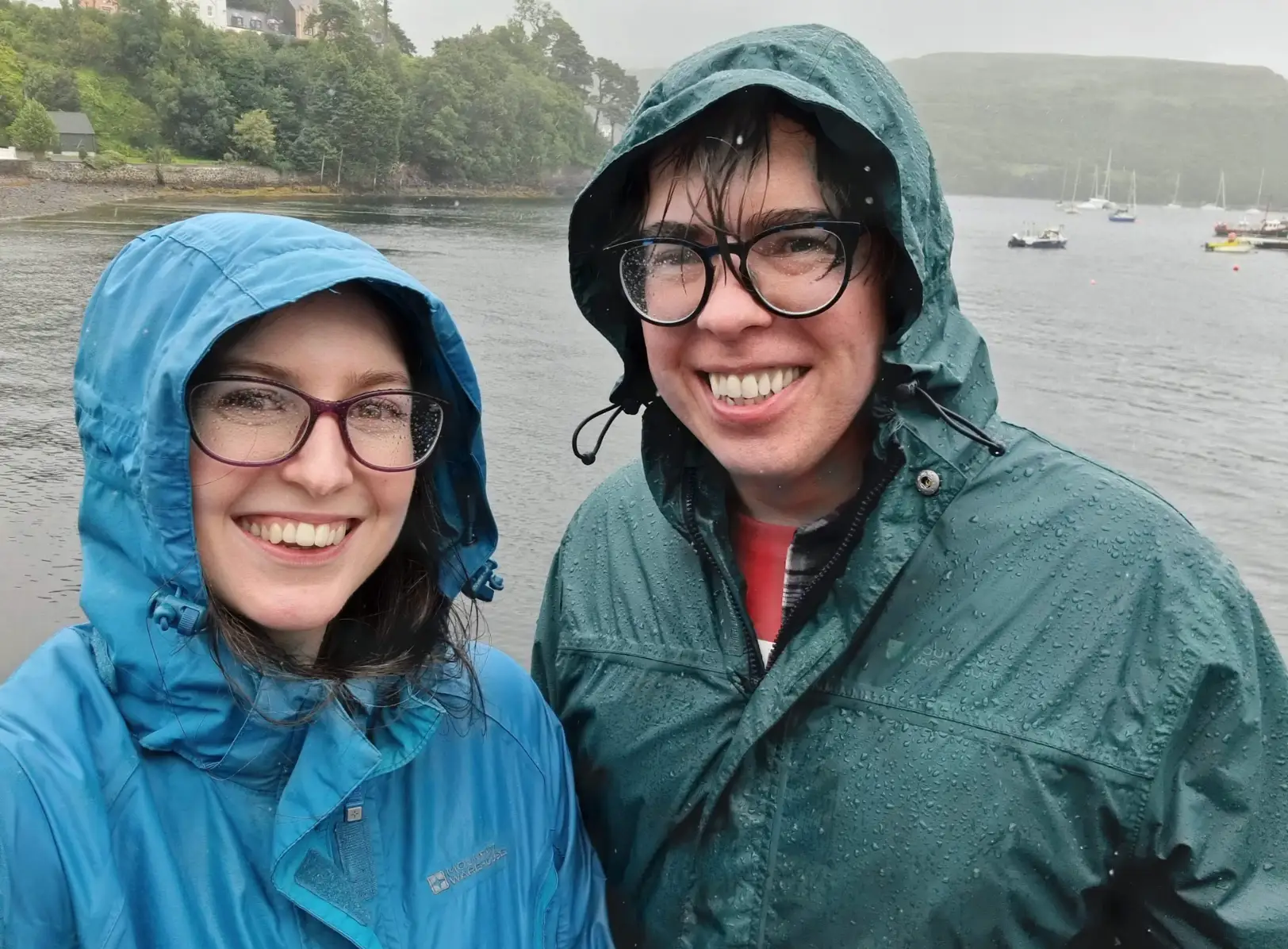 Two people wearing raincoats in Scotland, with a body of water and boats in the background.