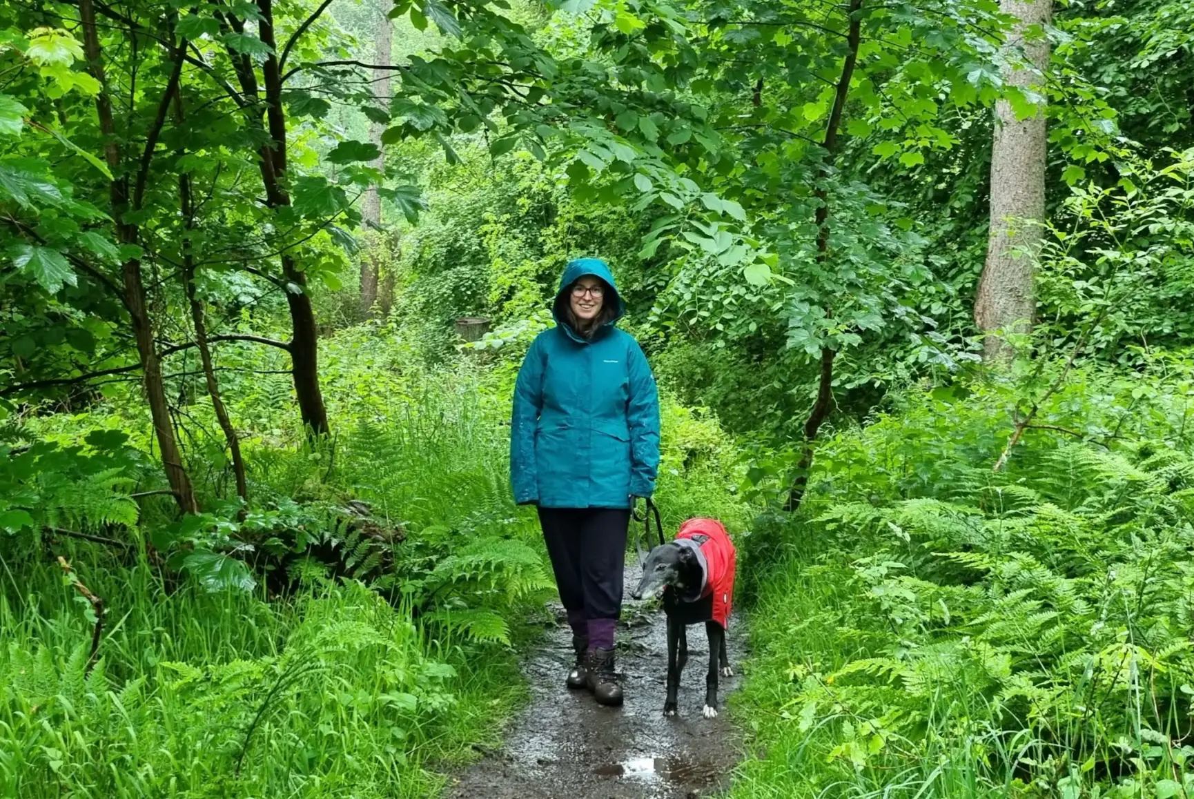 The writer and her dog, both wearing raincoats, walking down a muddy path amid trees in Scotland.