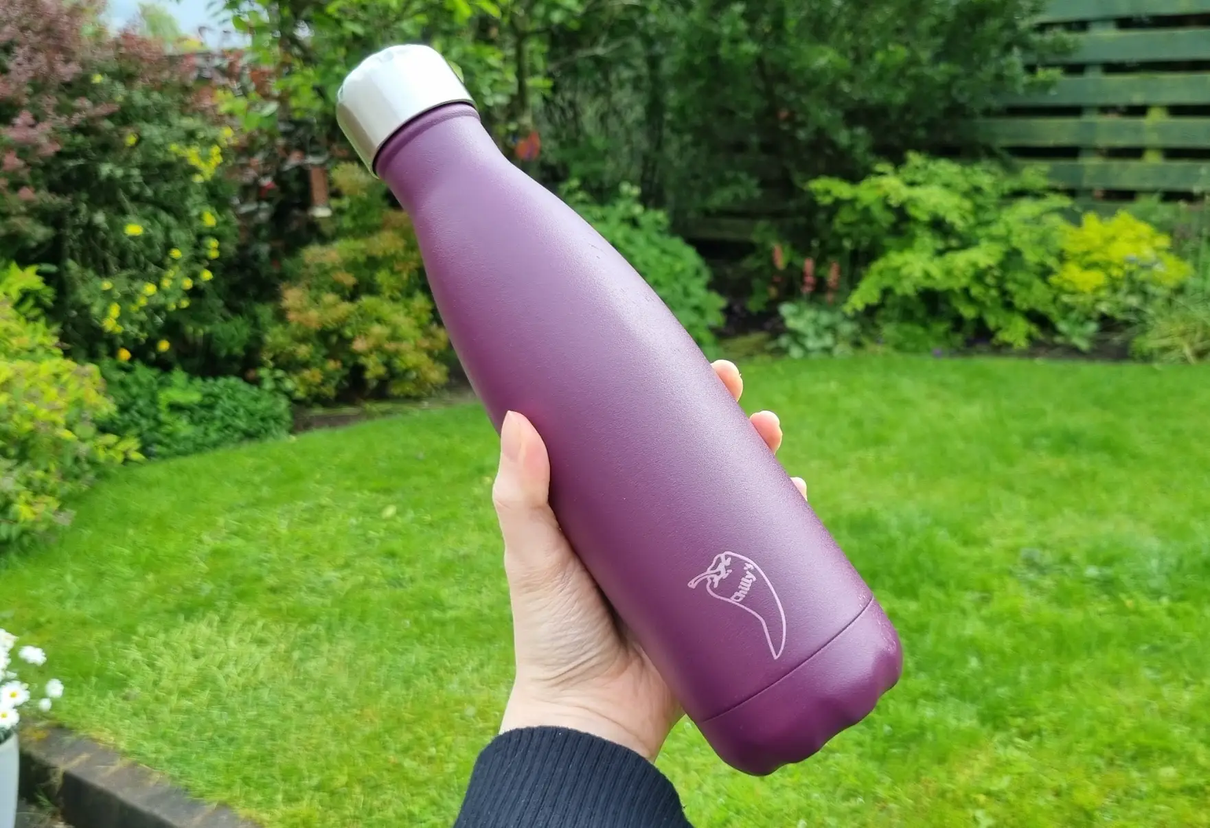 A hand holding up a purple reusable water bottle, with grass in the background.