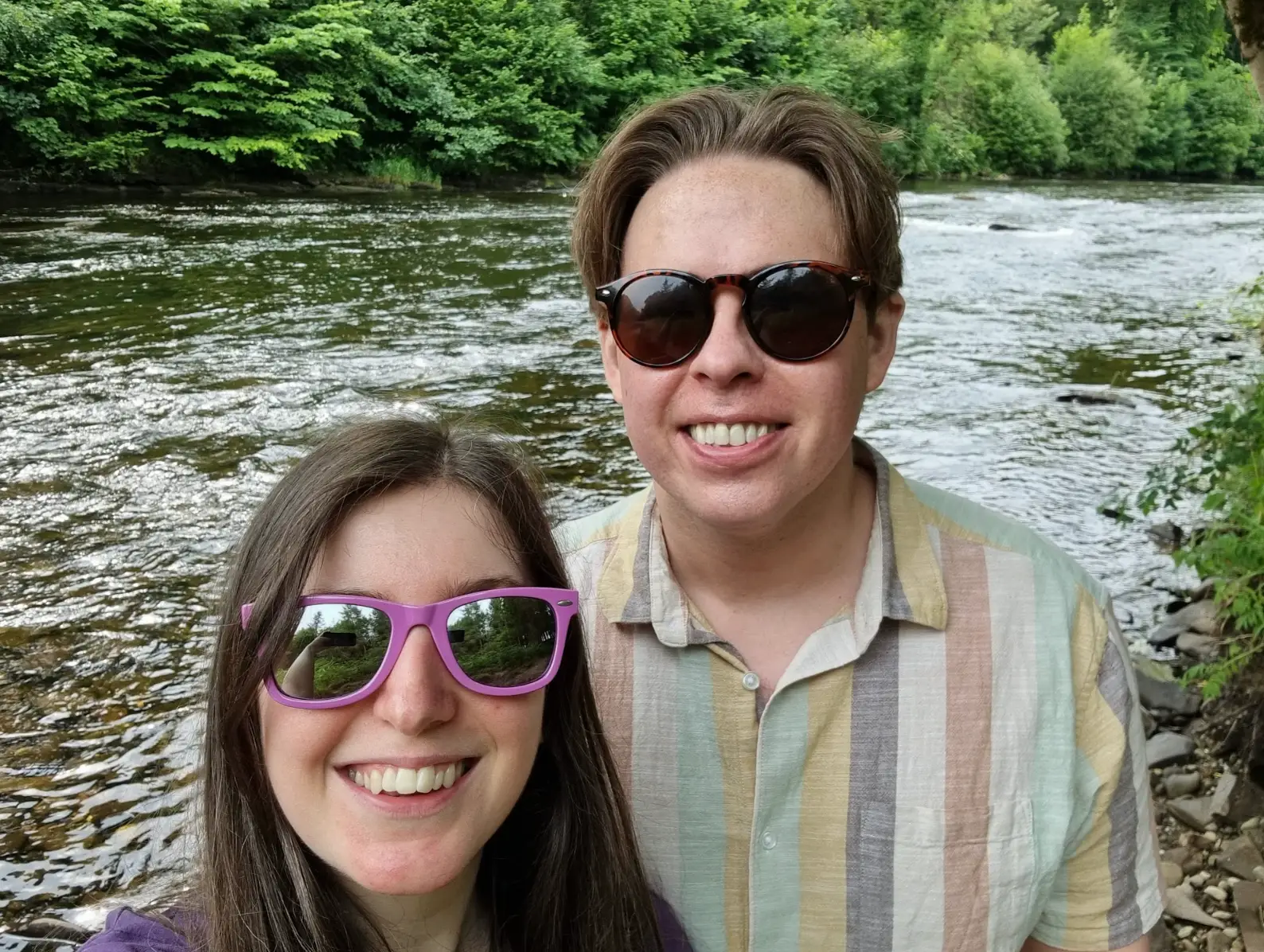 Two people wearing tinted sunglasses and standing in front of a body of water in Scotland.
