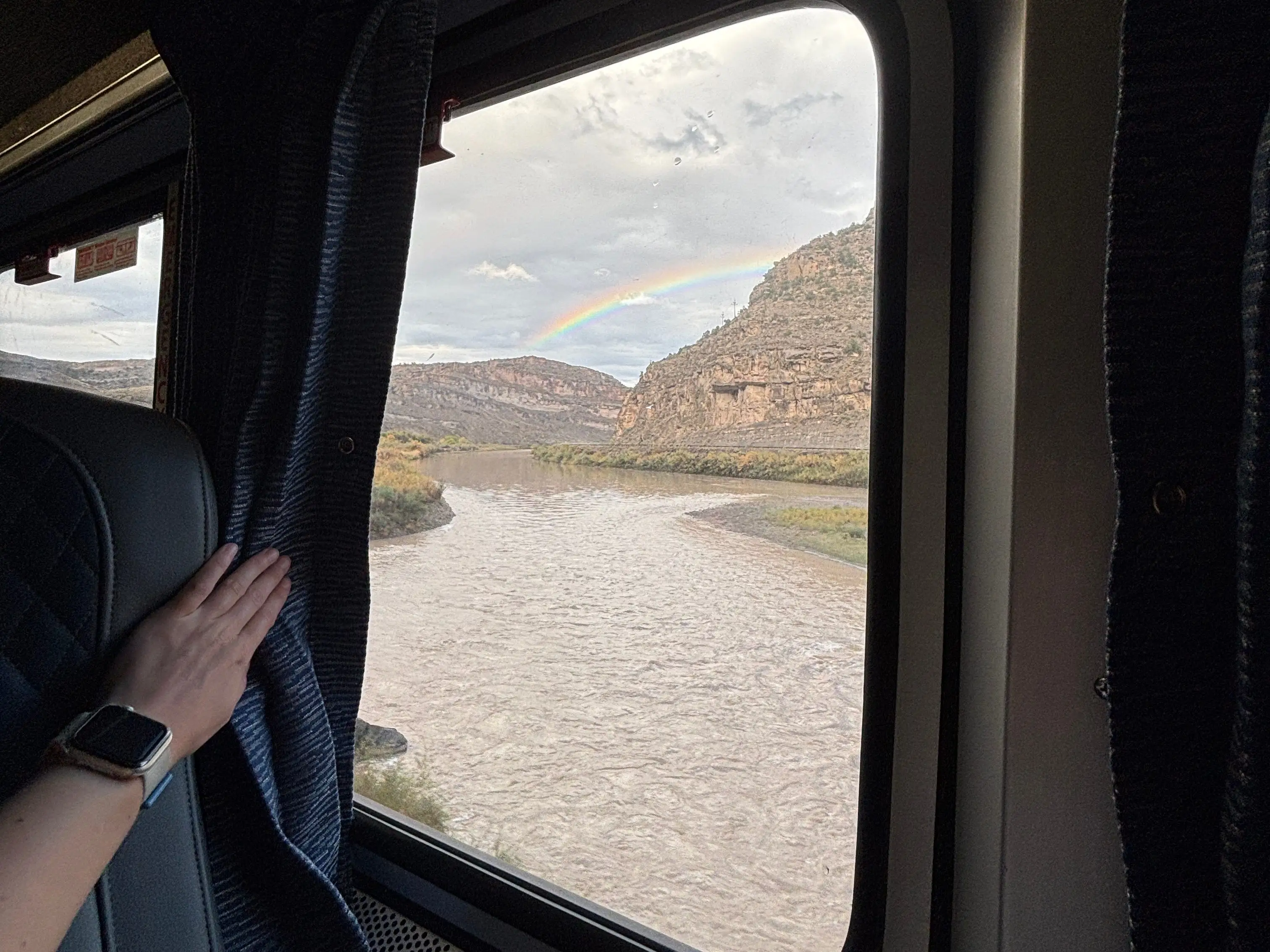 The window of an Amtrak train shows a rainbow appearing between two mountains.