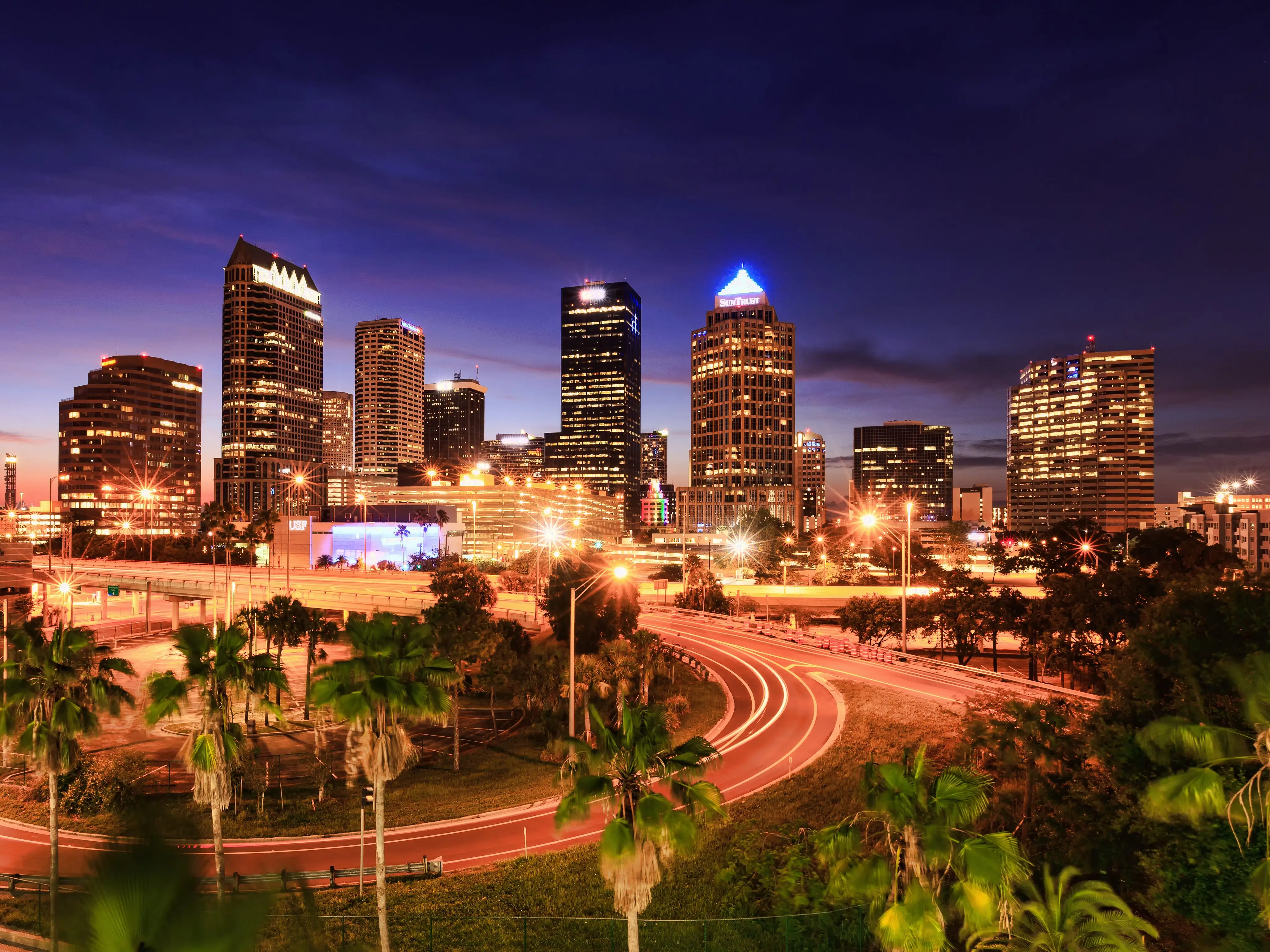 Downtown skyline glows at dusk with illuminated high-rises, palm trees, and curving highway light trails.
