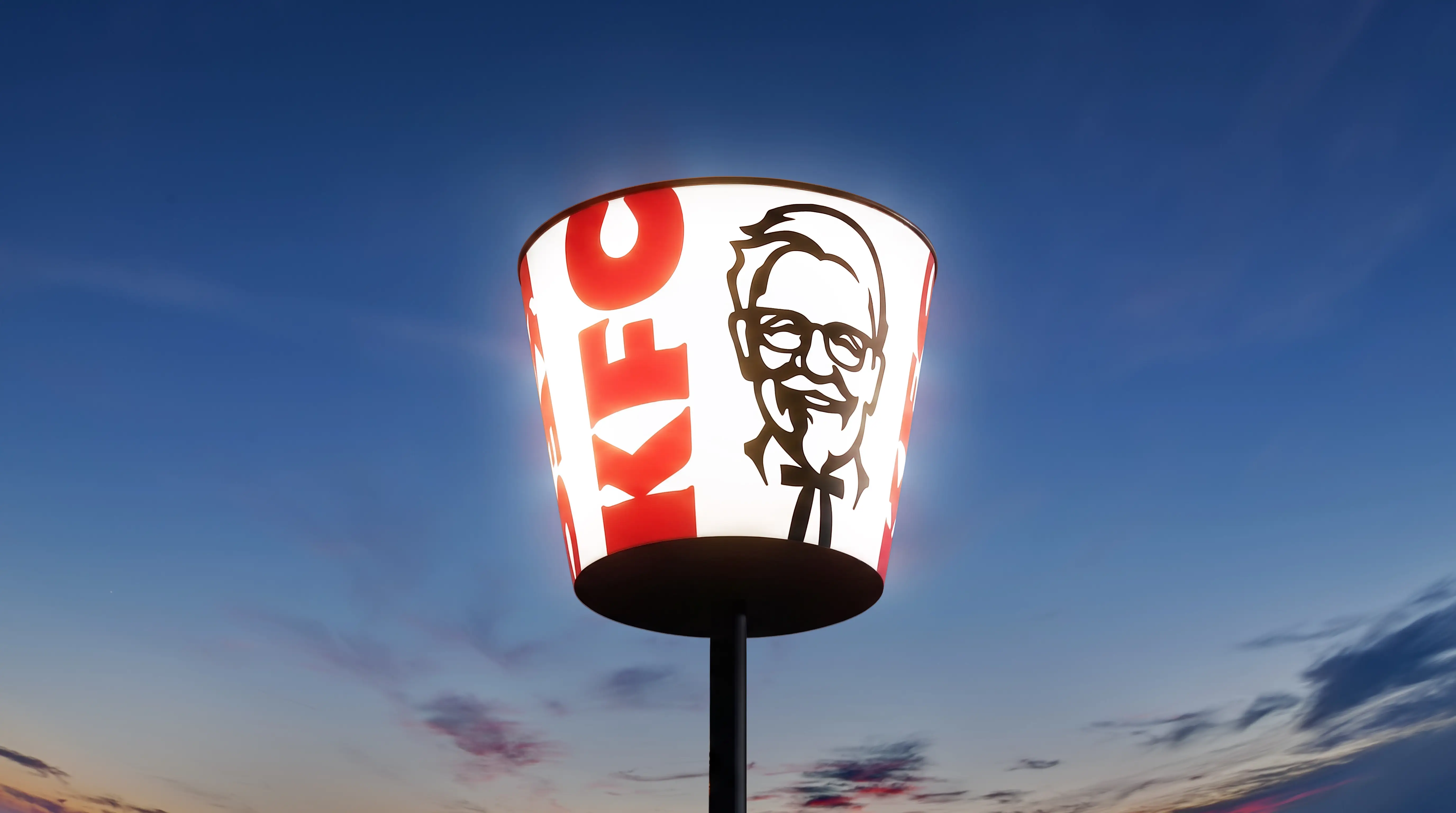 An illuminated KFC bucket sign in front of a dusky sky