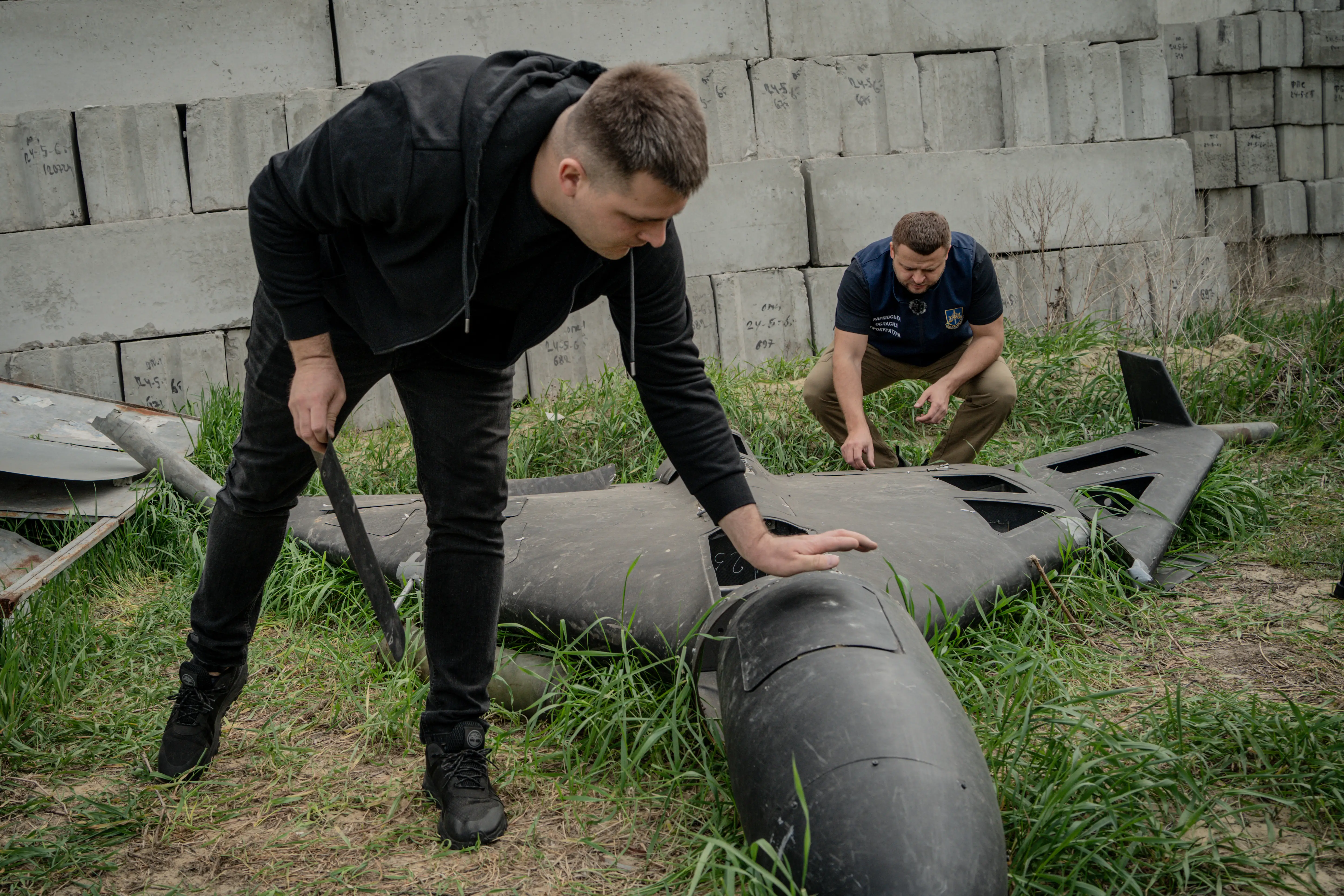 Two men lean over a large black drone on grass with concrete bricks stacked into a wall behind them