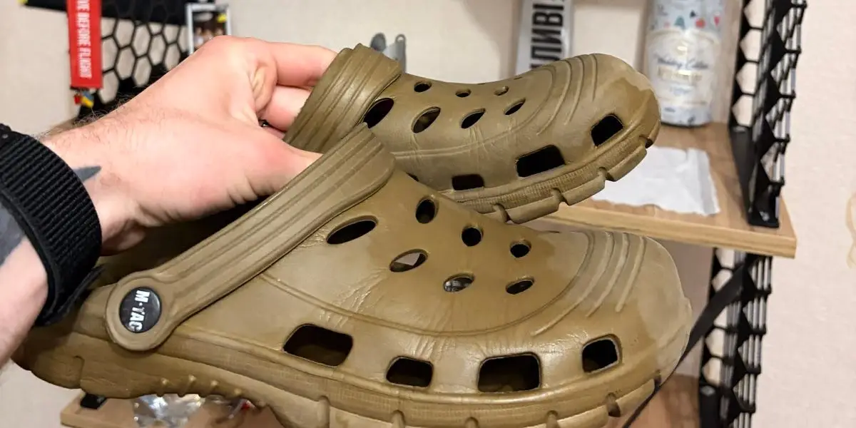 A hand holds up a pair of khaki rubber shoes with breathing holes in them against a set of wooden shelves that have a grey drone on them