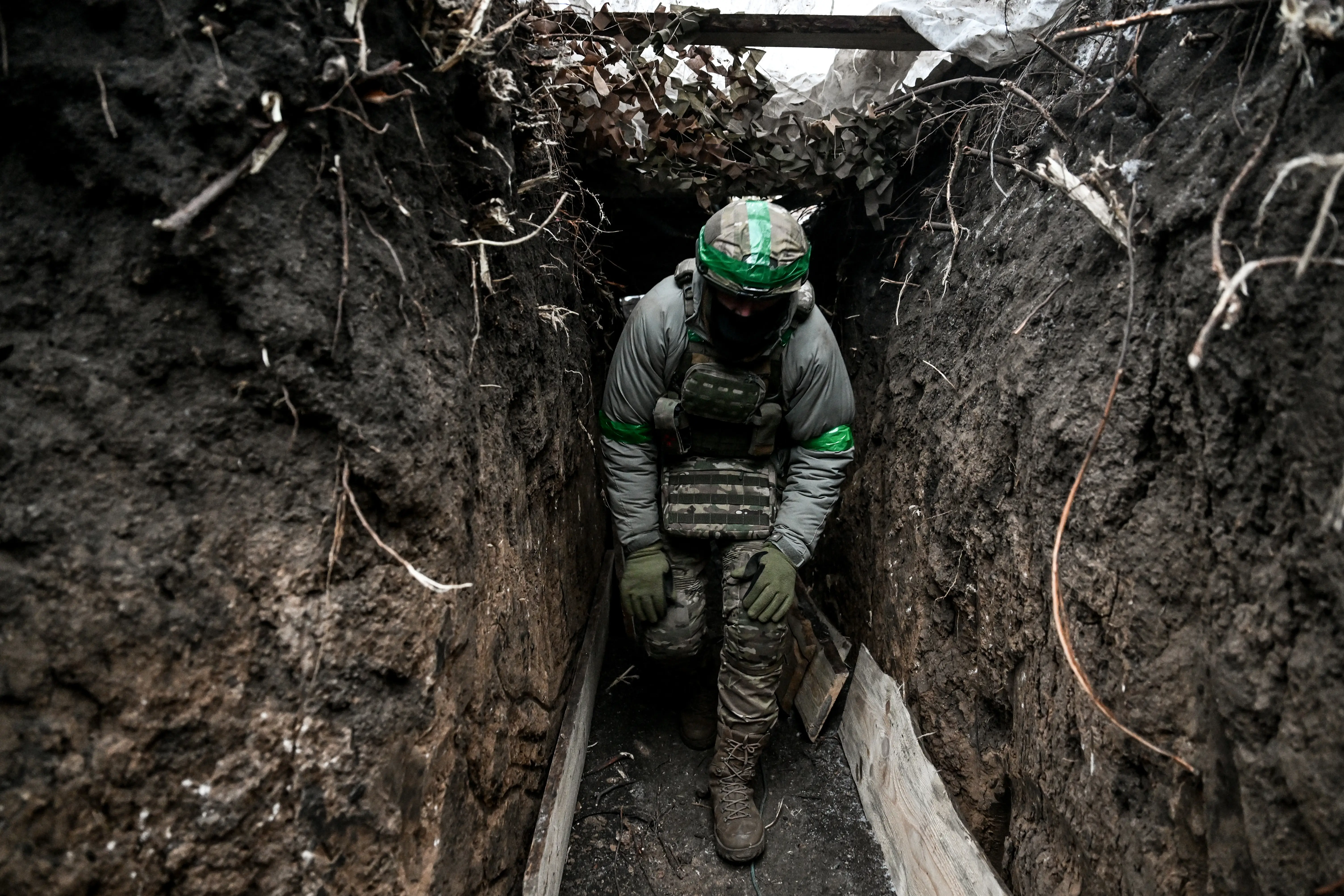 A man in camouflage gear and a helmet and boots walks bent over through a muddy trench
