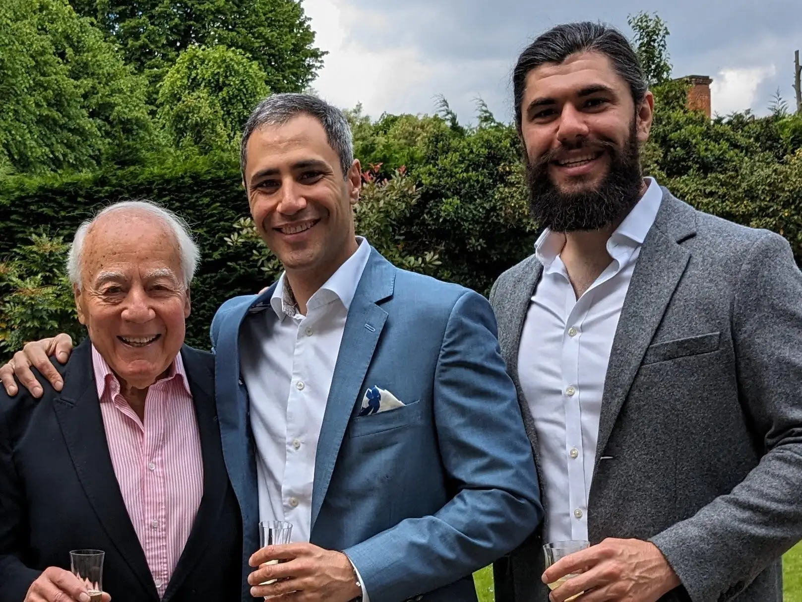 Three formally dressed people stand together outdoors in a garden, two holding champagne flutes.